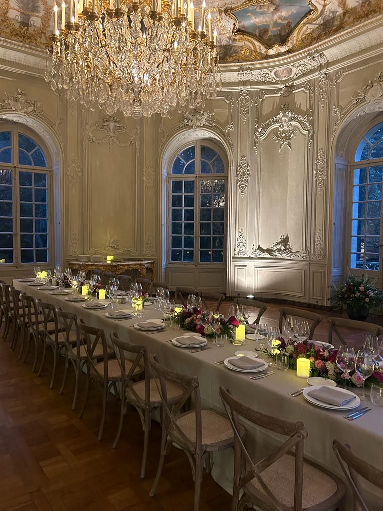 Elegant long dining table set with plates, wine glasses, floral arrangements, and glowing candles in a richly decorated room with large windows and a crystal chandelier.