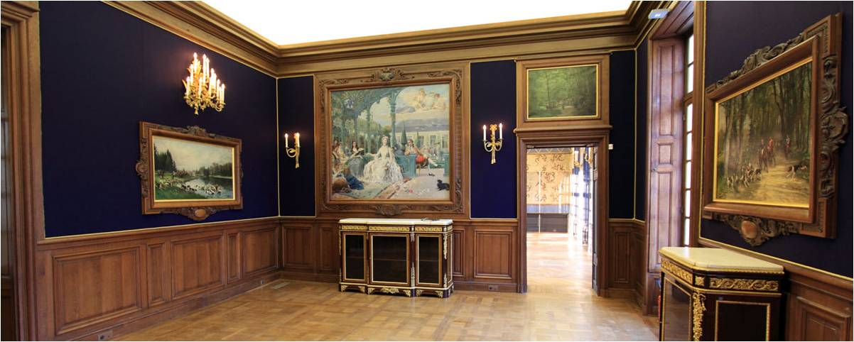 Interior of an elegant room with dark blue walls, wood paneling, framed classic paintings, chandeliers, and ornate furniture.
