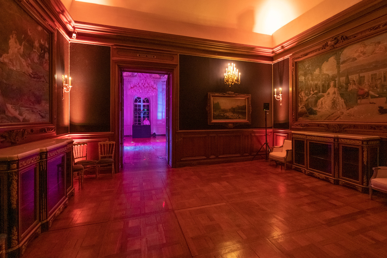 Ornate room with wooden flooring, antique chairs, paintings on dark walls, and warm lighting leading to a brightly lit adjacent room with a DJ.