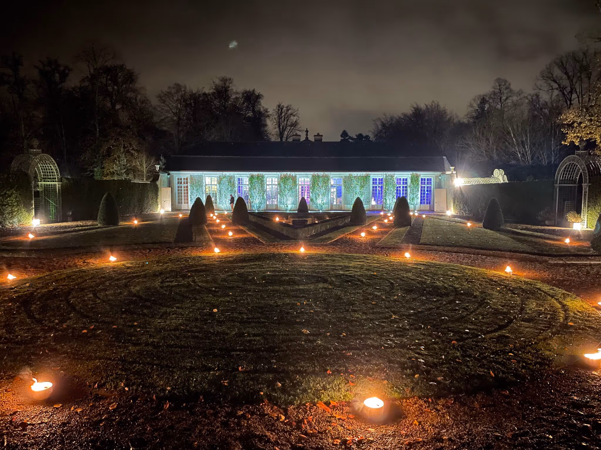 Night view of a garden with lit candles lining pathways leading to a well-lit building covered with climbing plants.