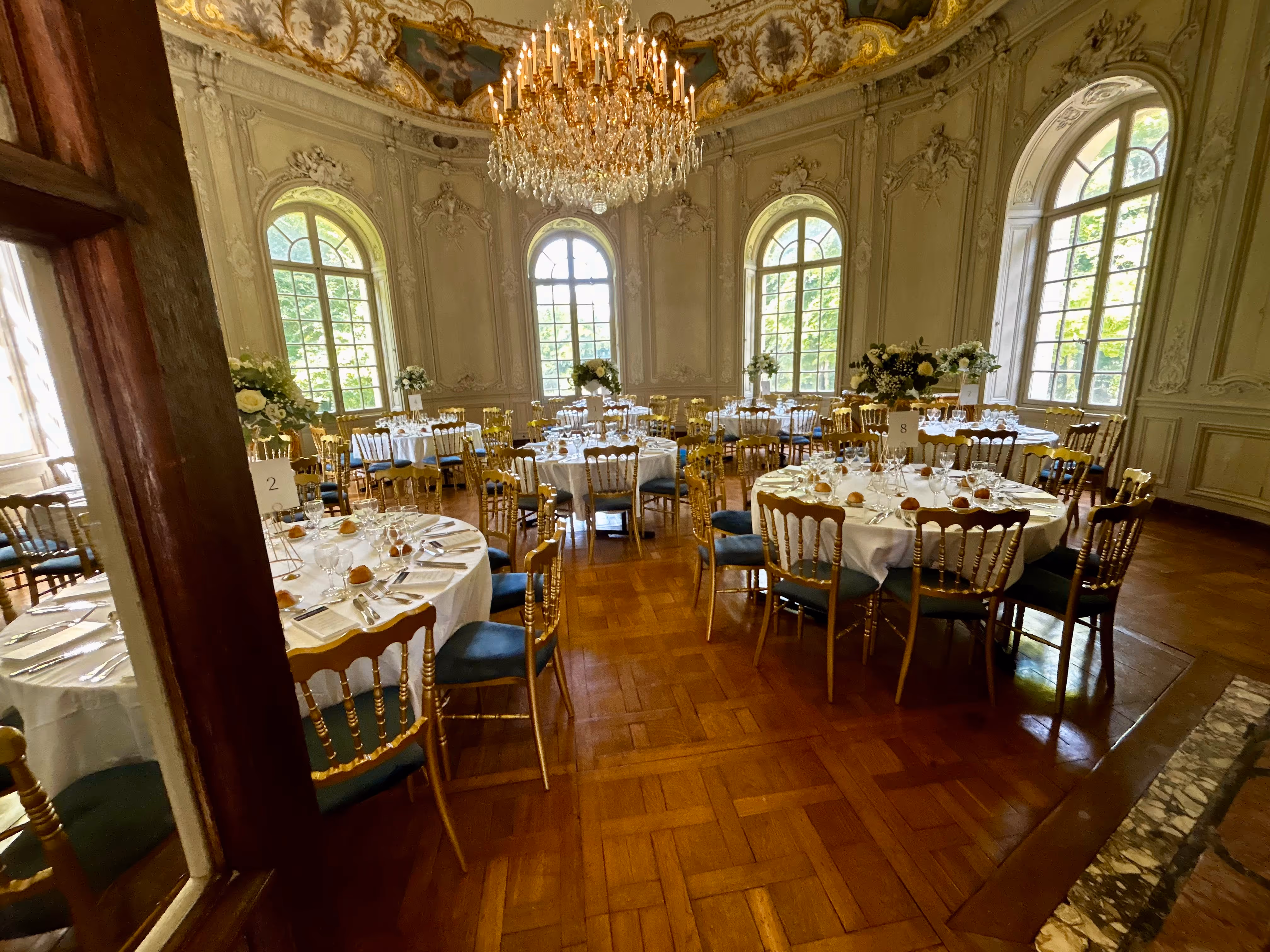 Elegant round banquet tables set with white tablecloths, gold chairs, glassware, and floral centerpieces under a large chandelier in a grand room with tall arched windows and ornate walls.