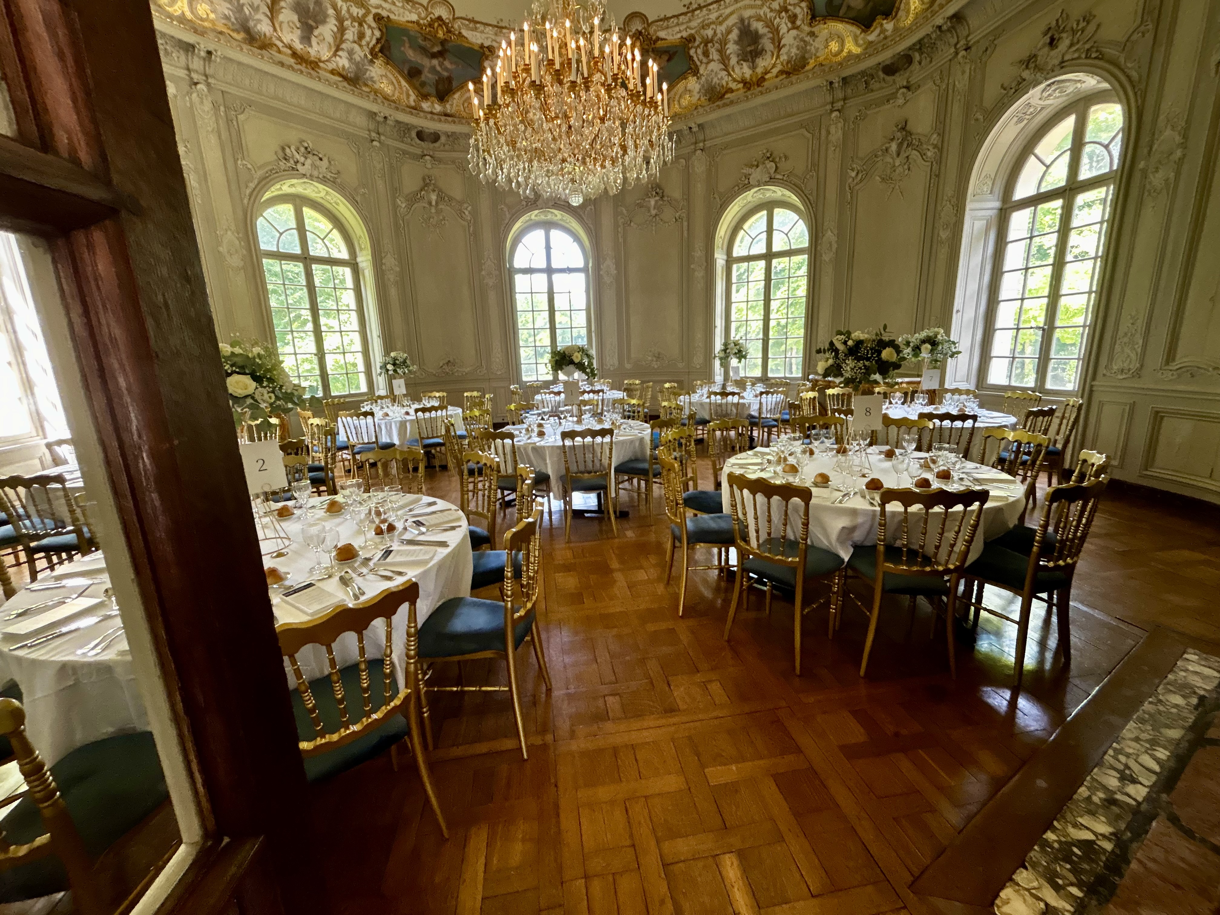 Elegant round banquet tables set with white tablecloths, gold chairs, glassware, and floral centerpieces under a large chandelier in a grand room with tall arched windows and ornate walls.