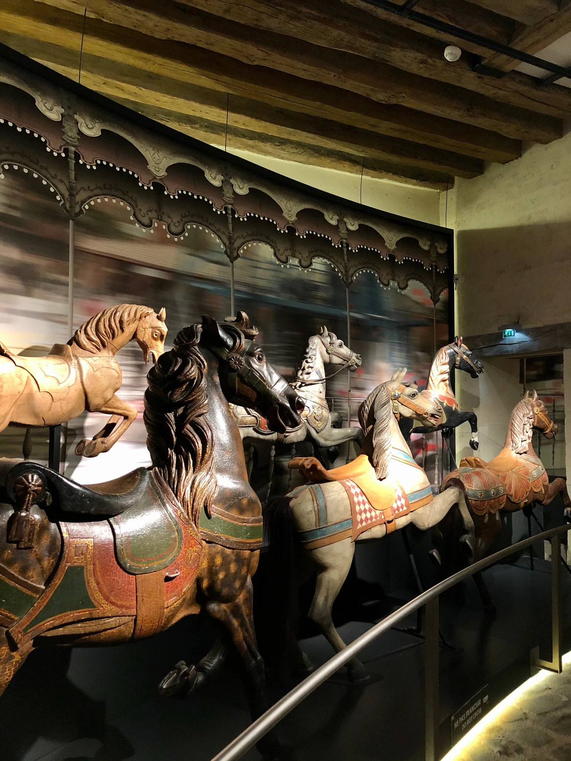 Exhibit of six vintage carousel horses on display in a dimly lit room with wooden beam ceiling.