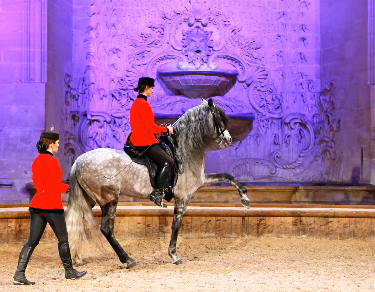 Woman in red jacket riding a gray horse lifting its front leg while another woman in a red jacket walks beside them in an ornate indoor arena.