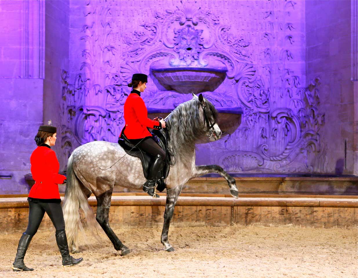 Woman in red jacket riding a gray horse lifting its front leg while another woman in a red jacket walks beside them in an ornate indoor arena.