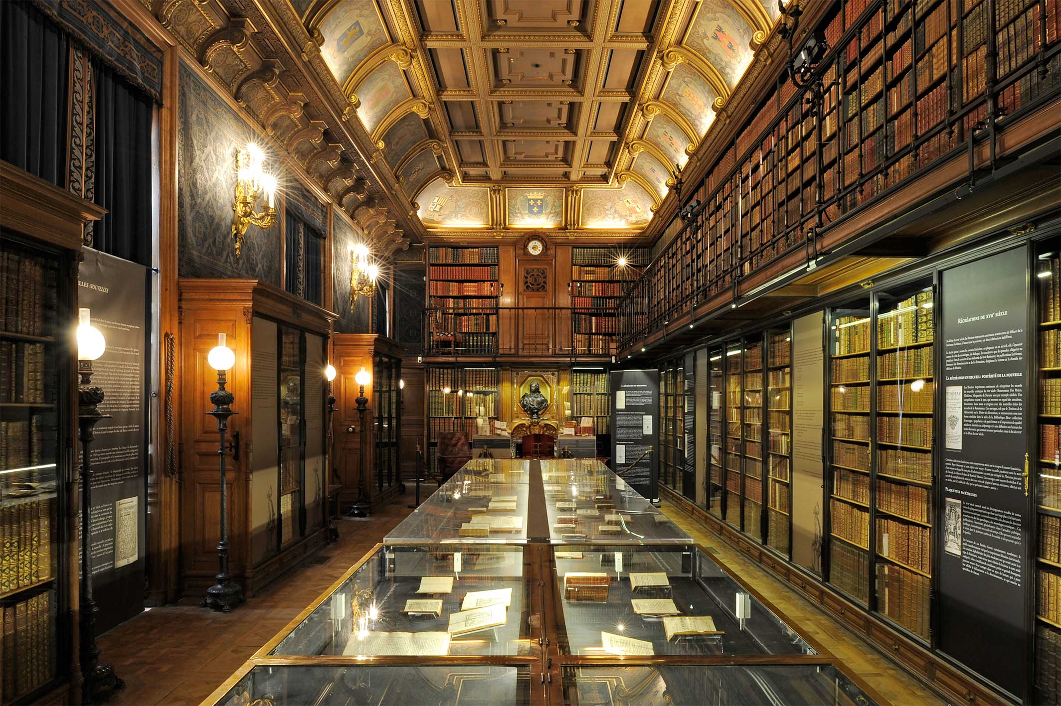 Ornate historic library room with tall wooden shelves filled with old books, a glass display table in the center holding open manuscripts, and a vaulted ceiling with paintings.