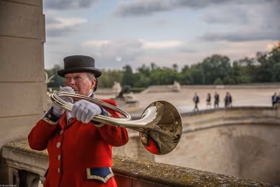 Man in a red coat and black top hat playing a brass horn outdoors near a stone railing with blurred people and trees in the background.