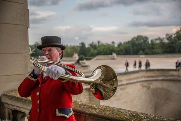 Man in a red coat and black top hat playing a brass horn outdoors near a stone railing with blurred people and trees in the background.