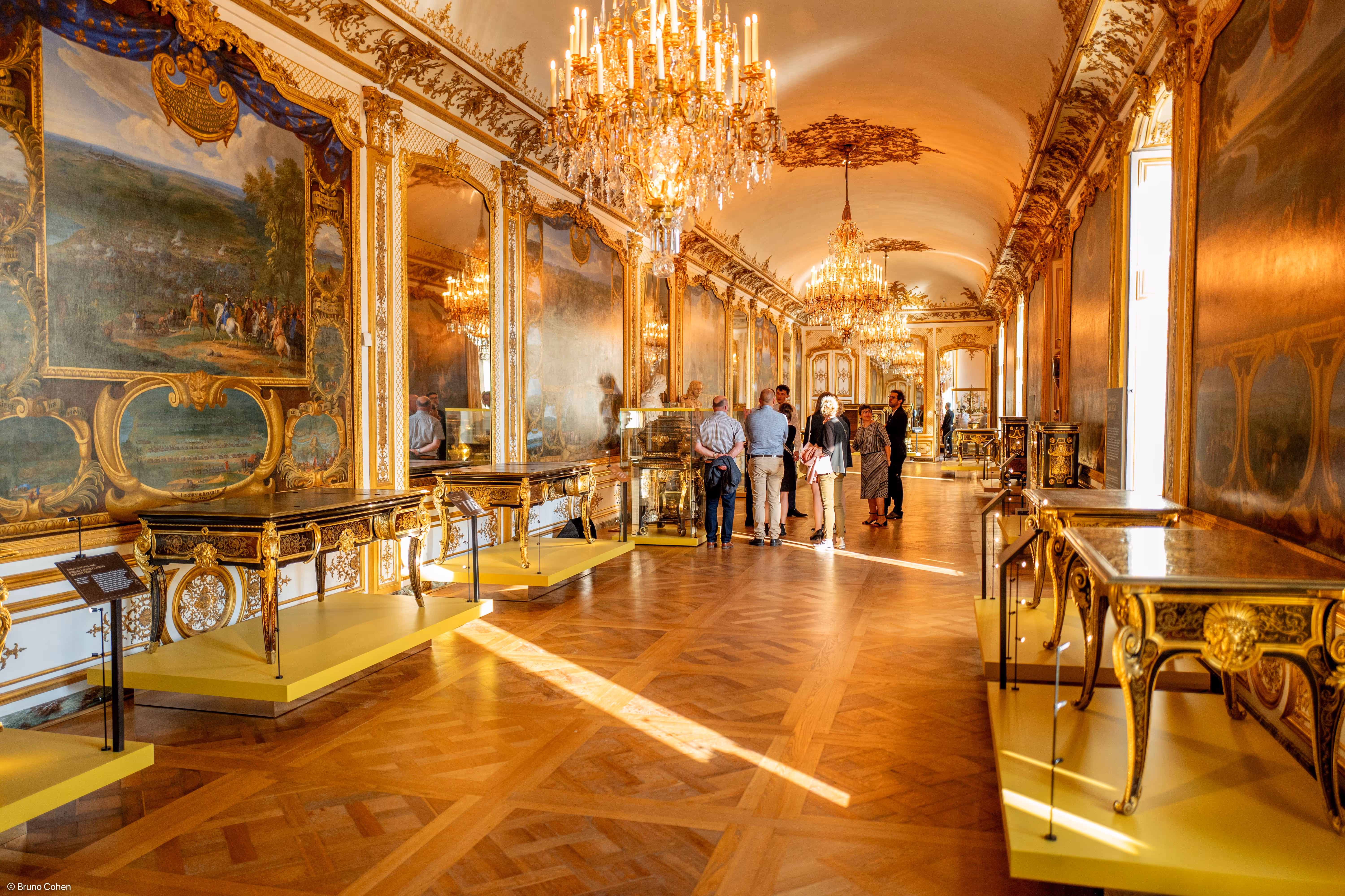 Visitors viewing ornate antique furniture and large gilded paintings in a grand, chandelier-lit museum gallery with parquet flooring.