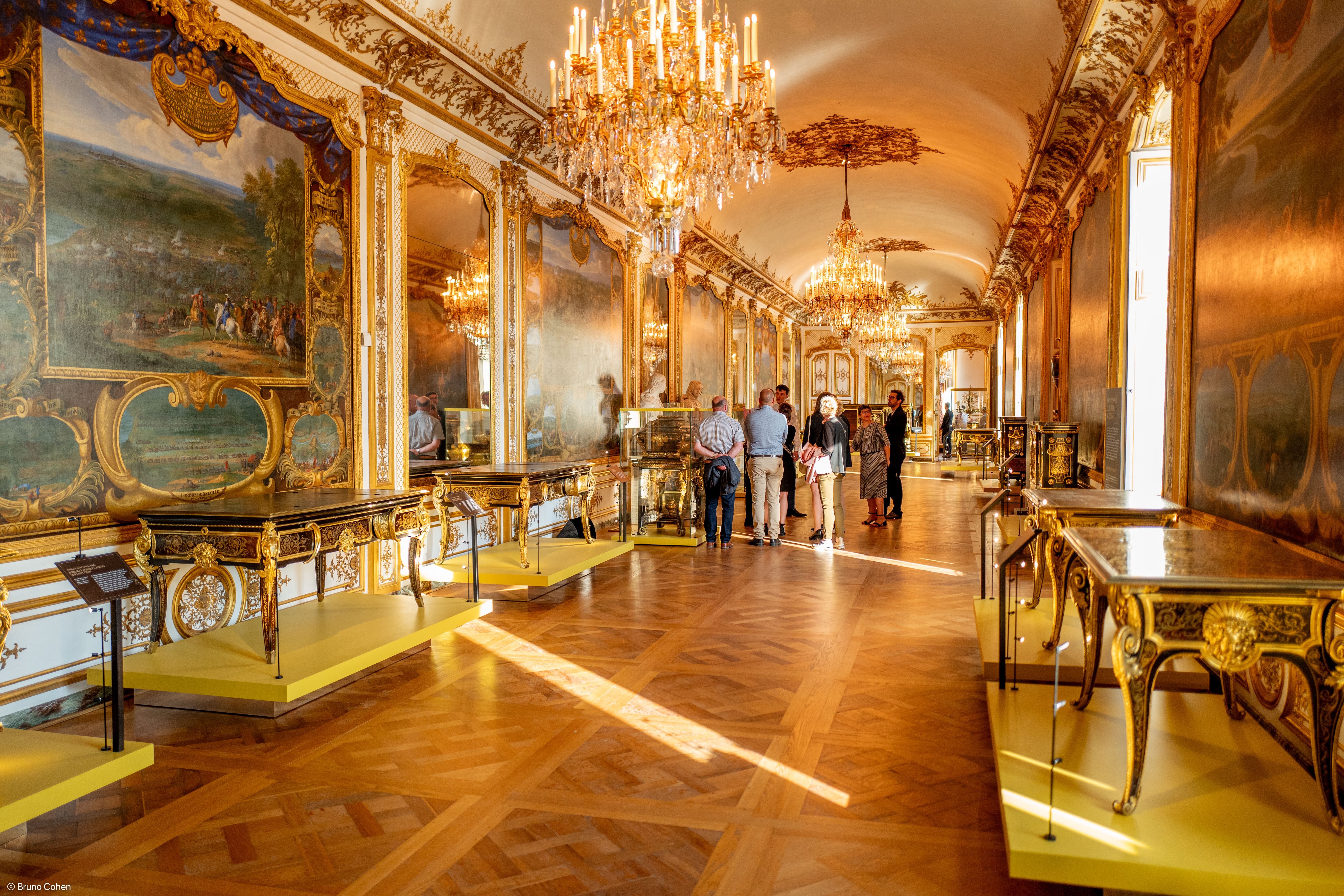 Visitors viewing ornate antique furniture and large gilded paintings in a grand, chandelier-lit museum gallery with parquet flooring.