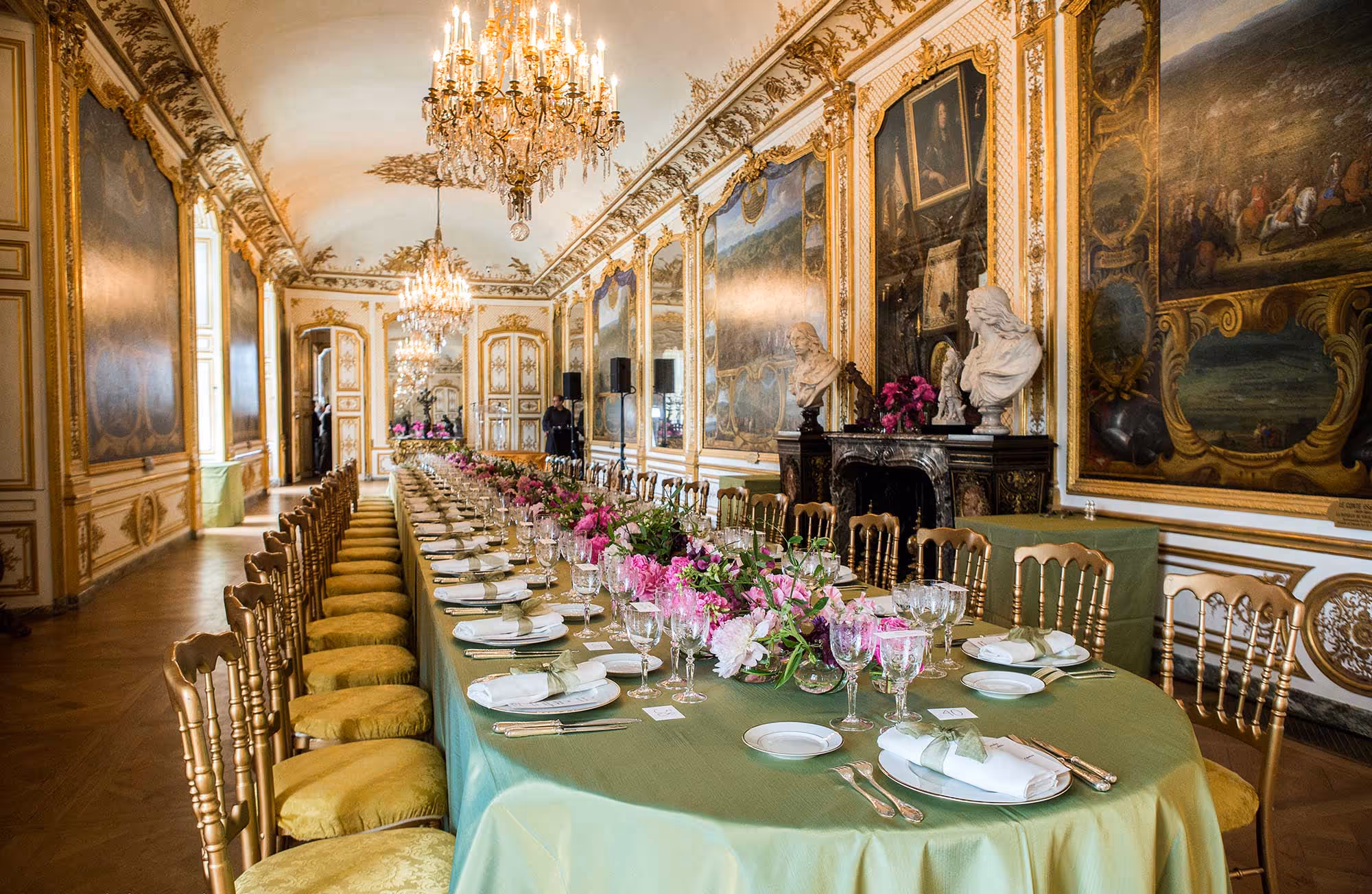 Long elegantly set dining table with green tablecloth, pink floral centerpieces, and crystal glassware in an ornate room with chandeliers and murals.