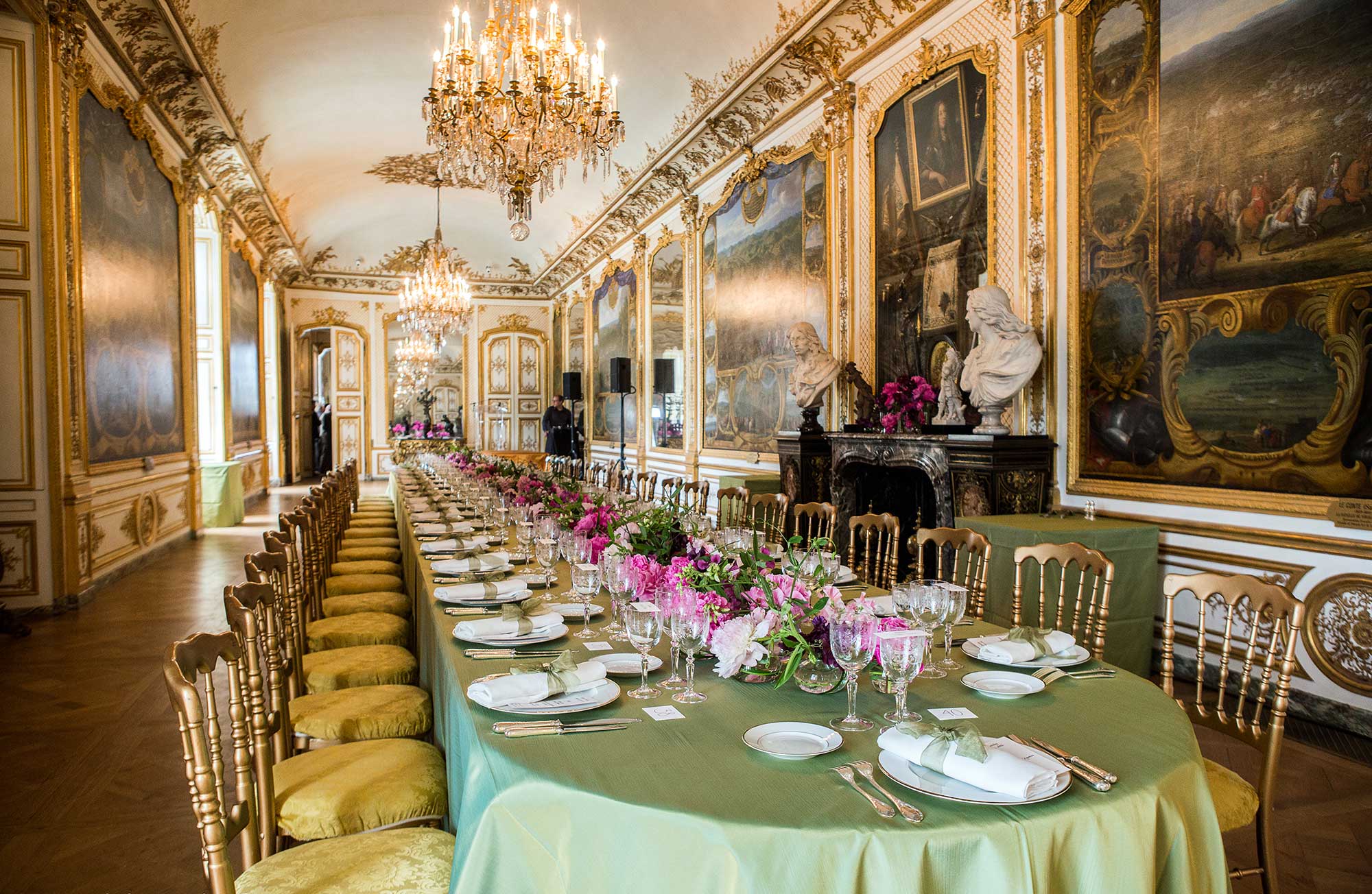 Long elegantly set dining table with green tablecloth, pink floral centerpieces, and crystal glassware in an ornate room with chandeliers and murals.