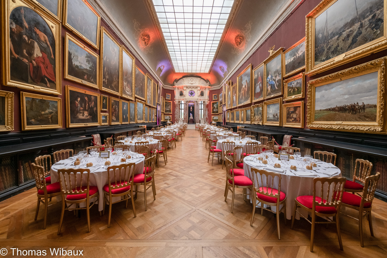 Elegant dining hall with round tables set for a banquet, surrounded by walls covered with large framed paintings.