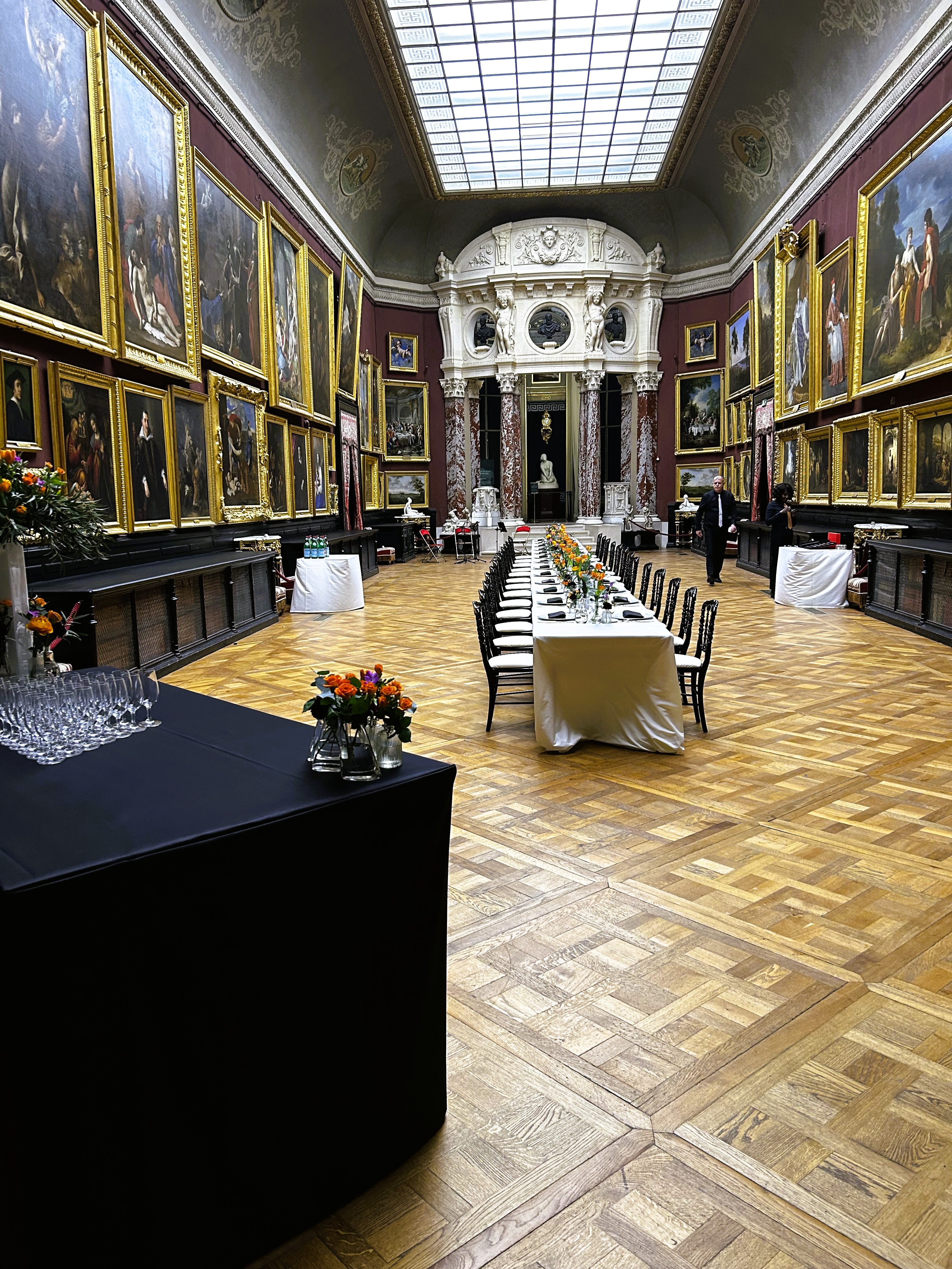 Elegant banquet setup in a grand hall with parquet flooring, ornate paintings, and a large skylight ceiling.
