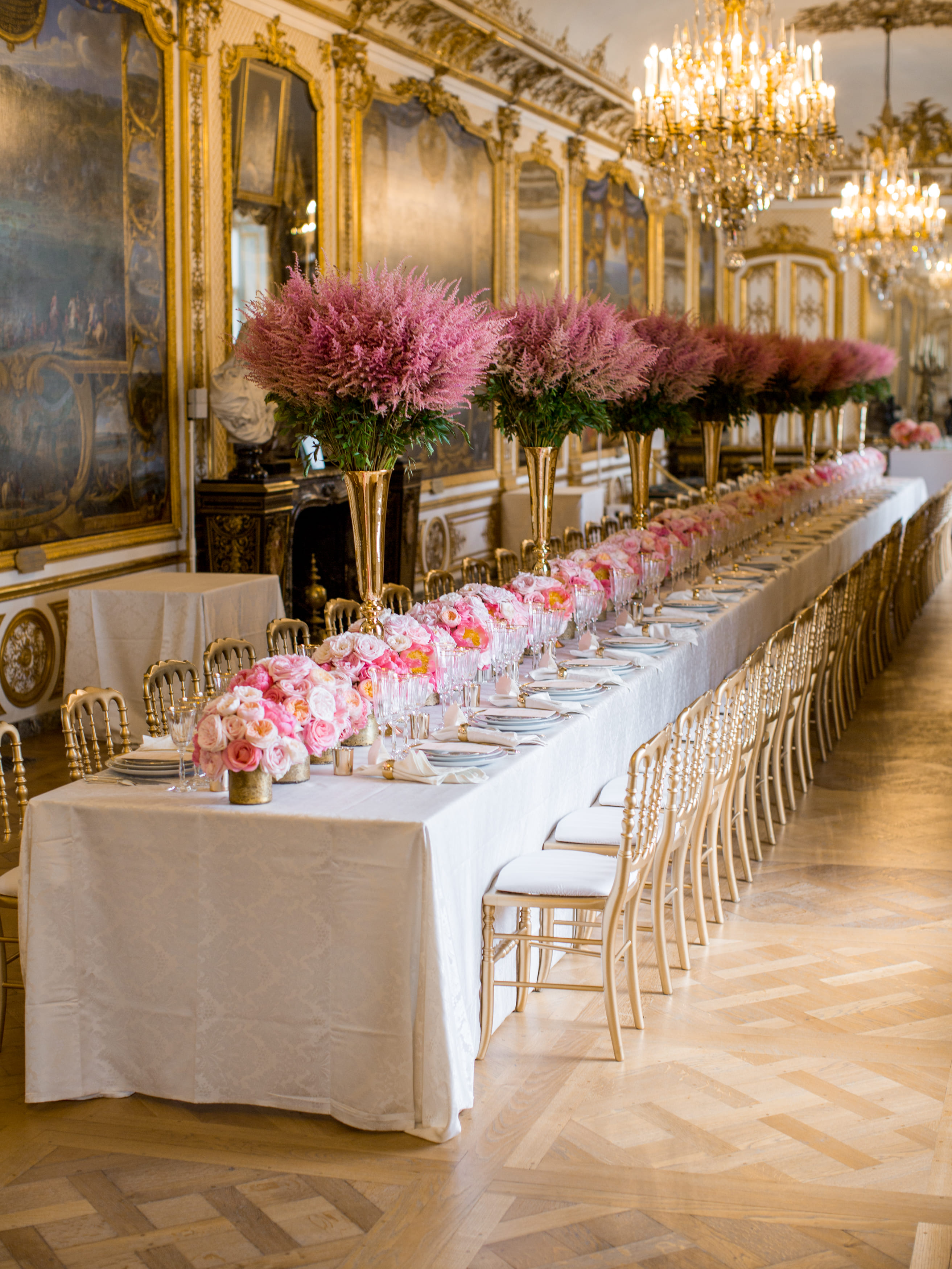Long elegant dining table in a grand room, decorated with tall pink floral centerpieces and gold chairs.