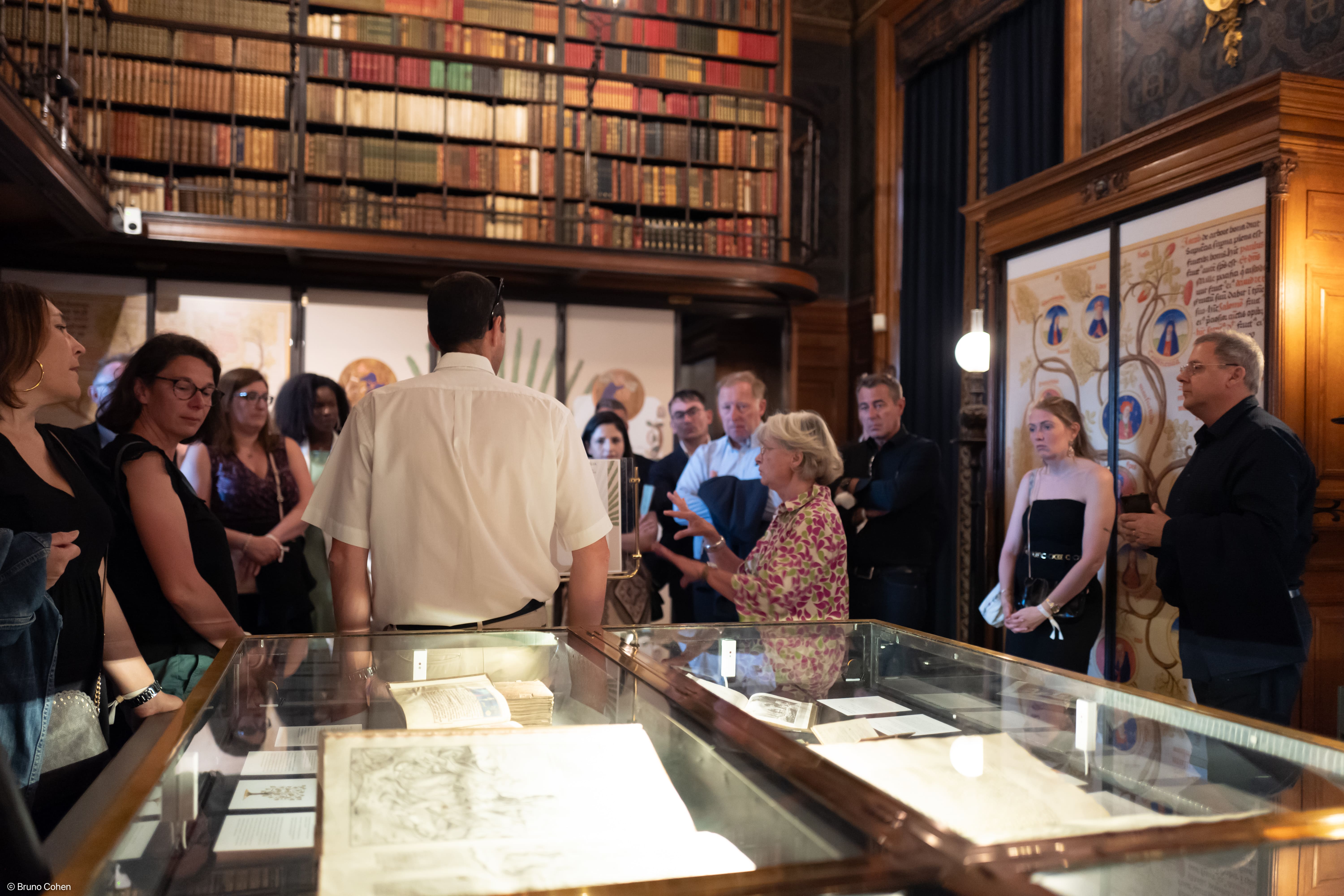 Group of people attentively listening to a guide inside a historic library with glass display cases and shelves filled with books.