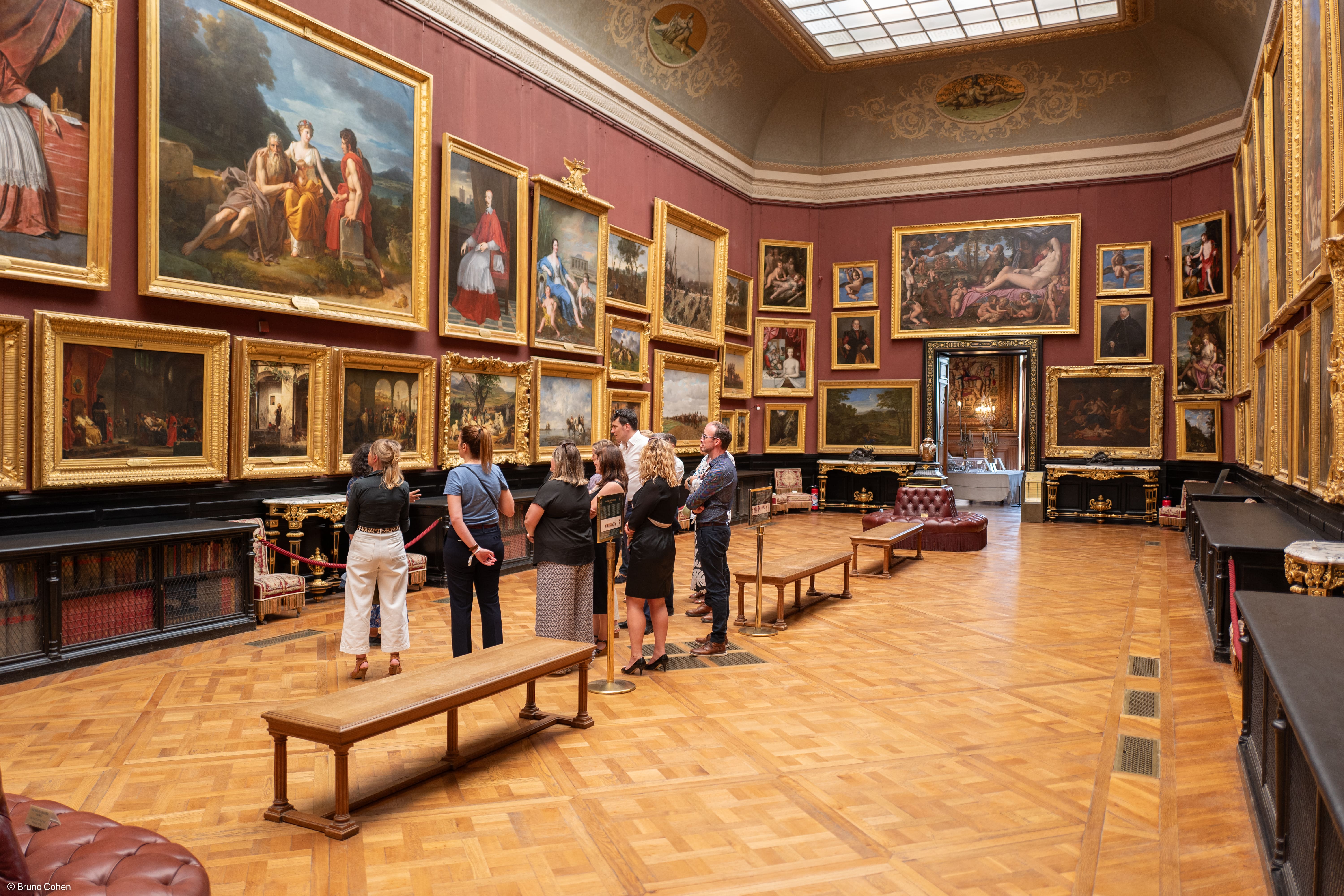 Group of people observing large framed classical paintings in an ornate museum gallery with wooden parquet floors and high ceiling.