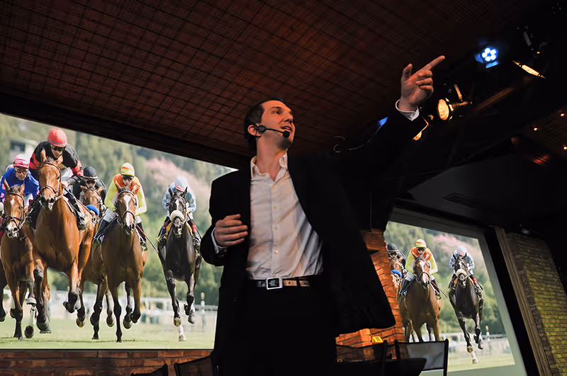 Man in a suit with a headset microphone gesturing and speaking in front of large screens showing horse racing footage.