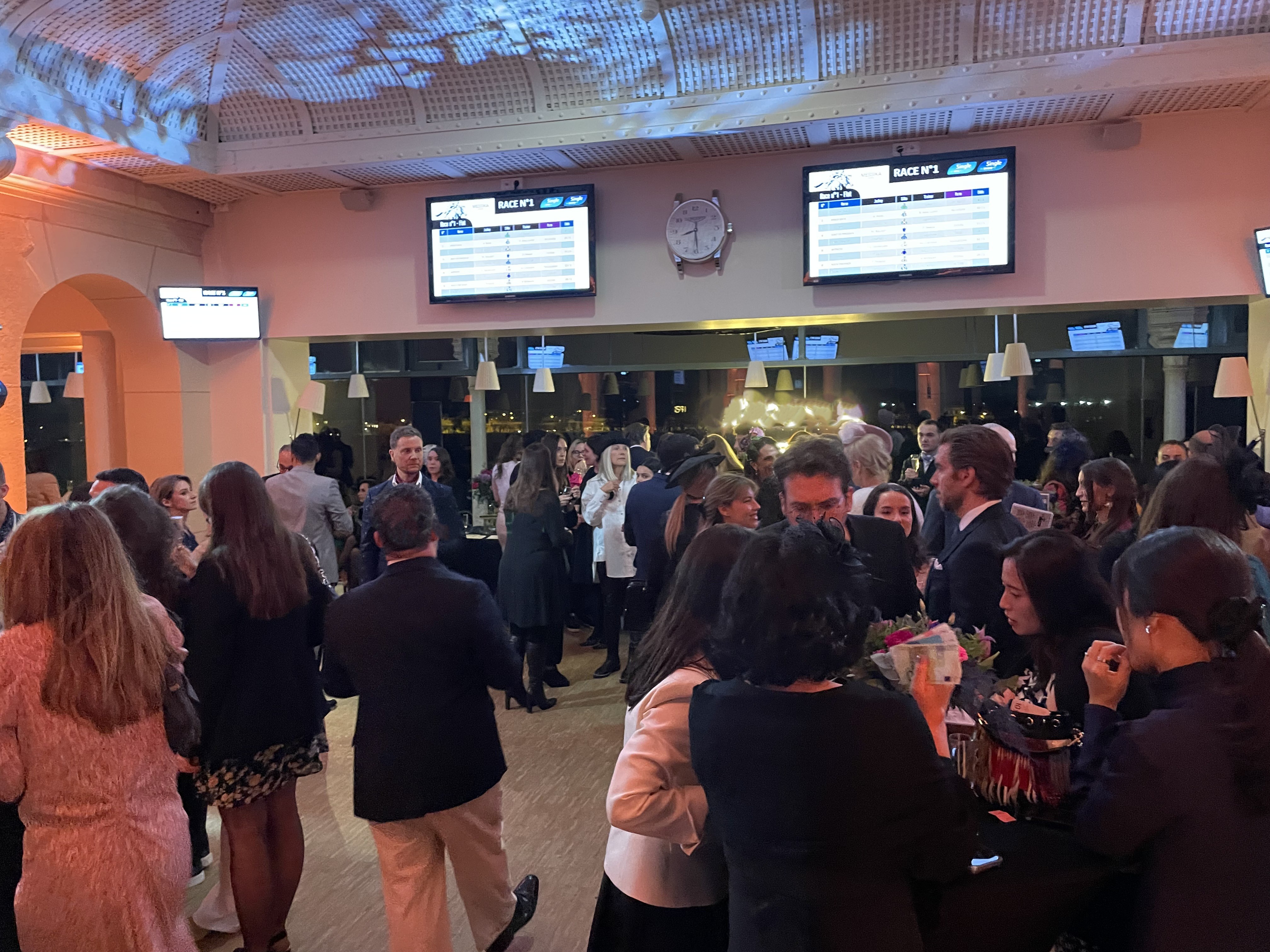 Crowd of people socializing indoors under a high ceiling with race information screens and a clock on the wall.