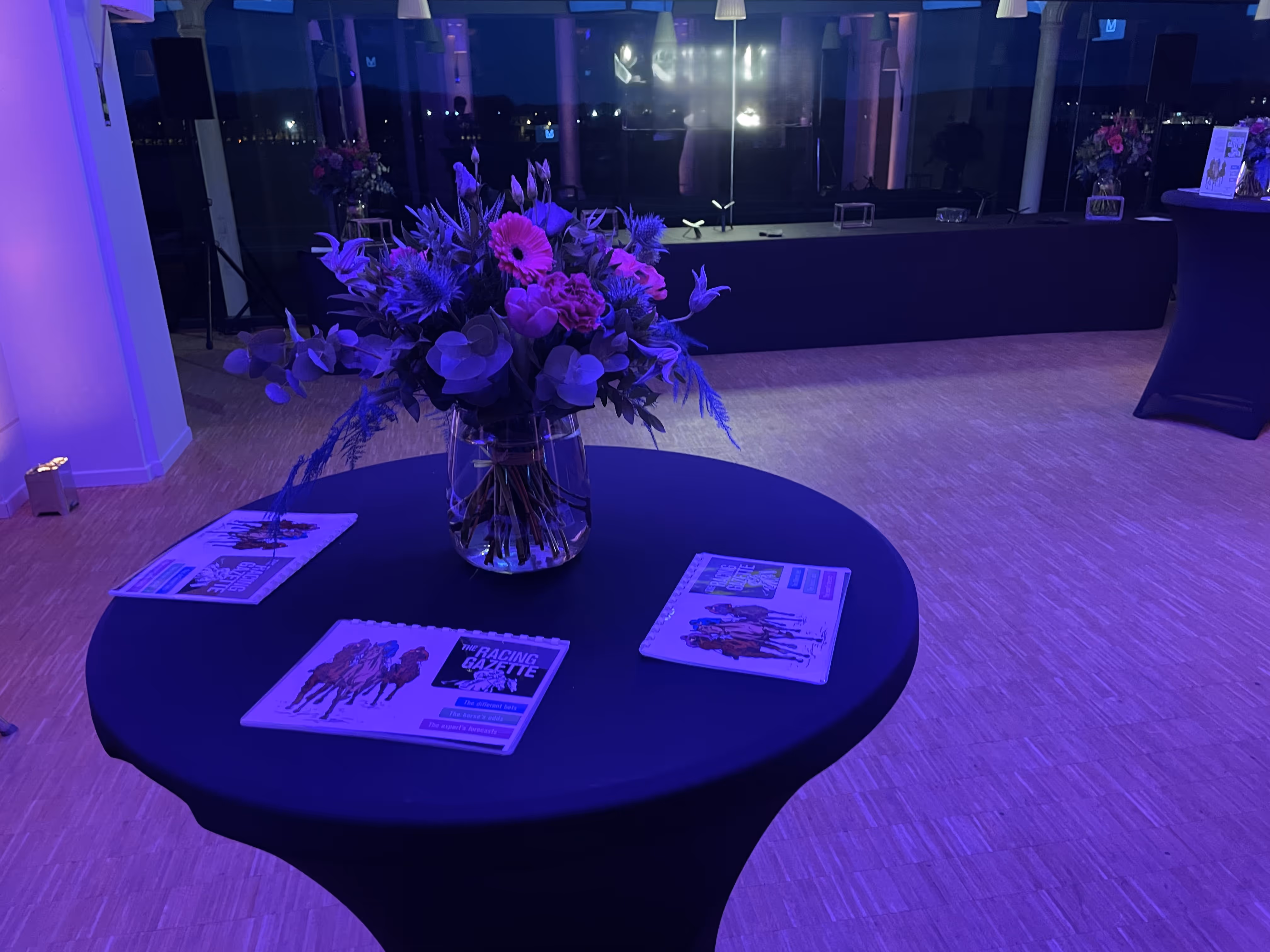 A round table with a dark cloth holds a glass vase of flowers and three copies of The Racing Gazette magazine, in a dimly lit room with blue lighting.