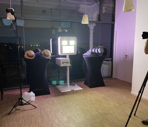 Indoor photo booth setup with two tables holding various hats and a bright light panel in the center, with a photographer partially visible on the right.