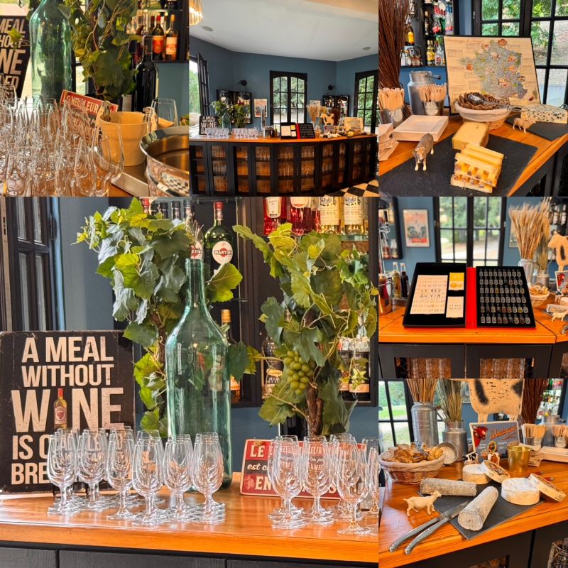 Interior of a wine tasting room with glasses on wooden tables, grapevine decorations, cheese displays, and a sign reading 'A meal without wine is called breakfast.'