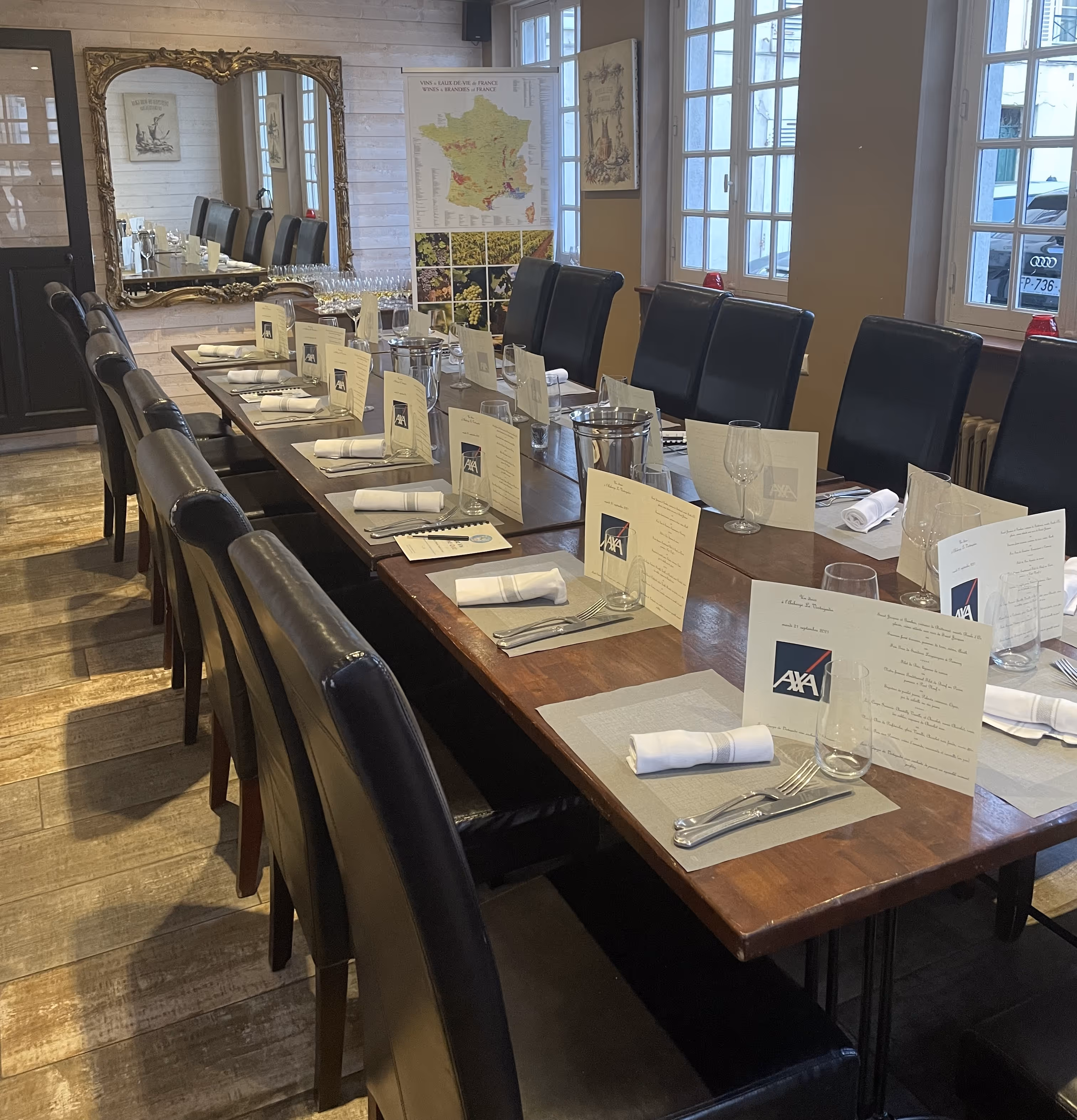 Long dining table set with menus, napkins, cutlery, and glasses in a warmly lit room with large windows and a decorative mirror.