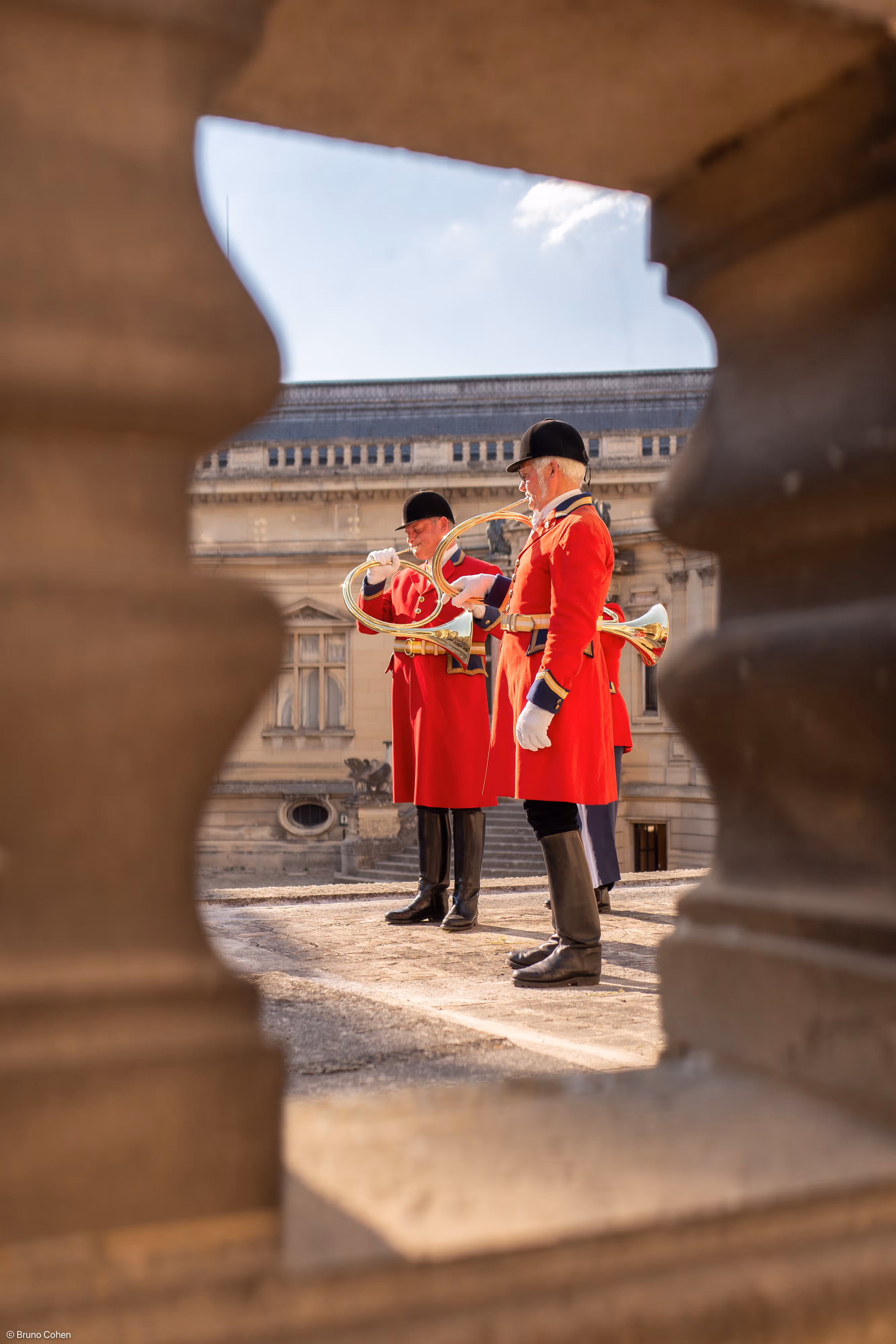 Three men in red ceremonial uniforms playing brass hunting horns, framed by stone balusters.