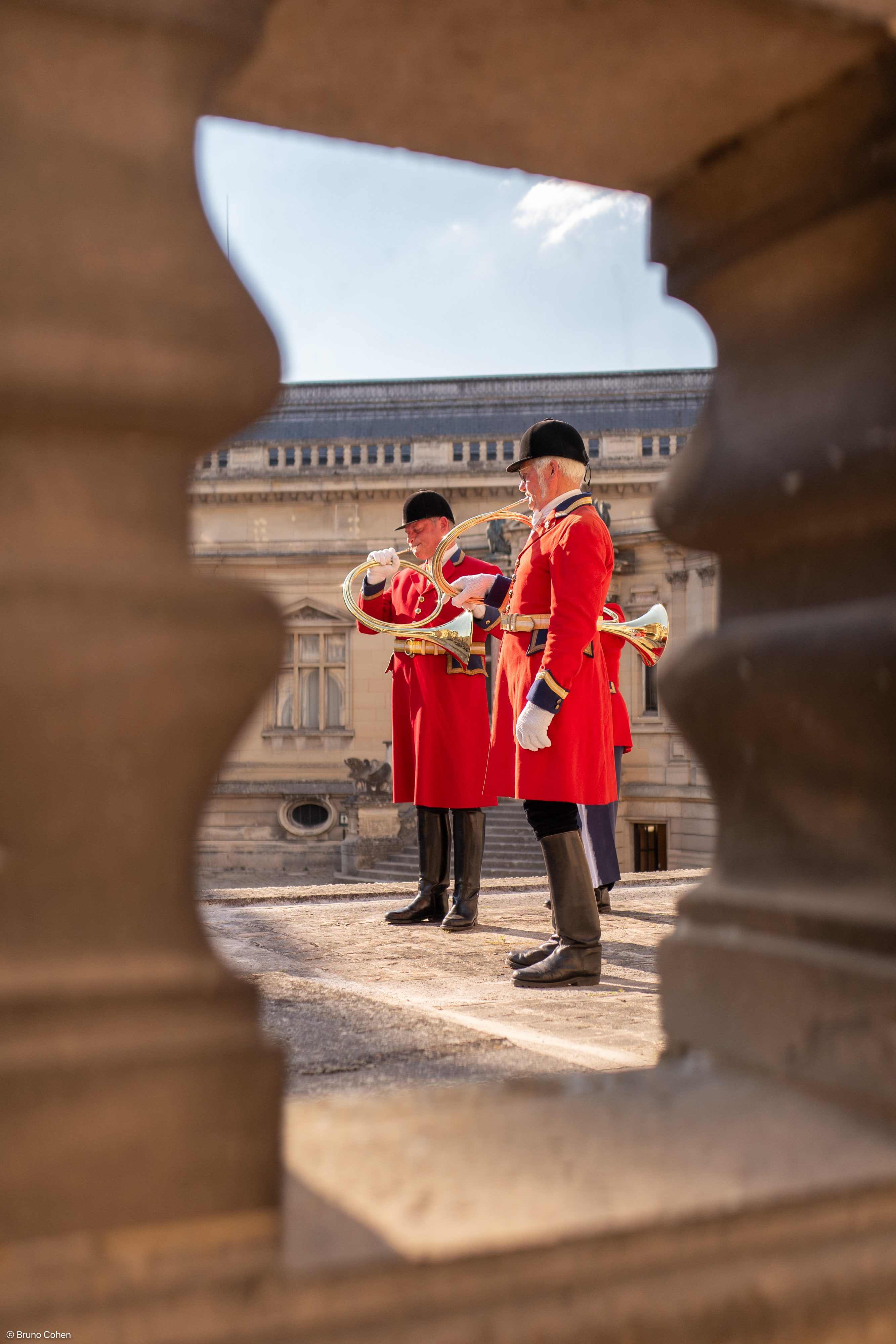 Three men in red ceremonial uniforms playing brass hunting horns, framed by stone balusters.