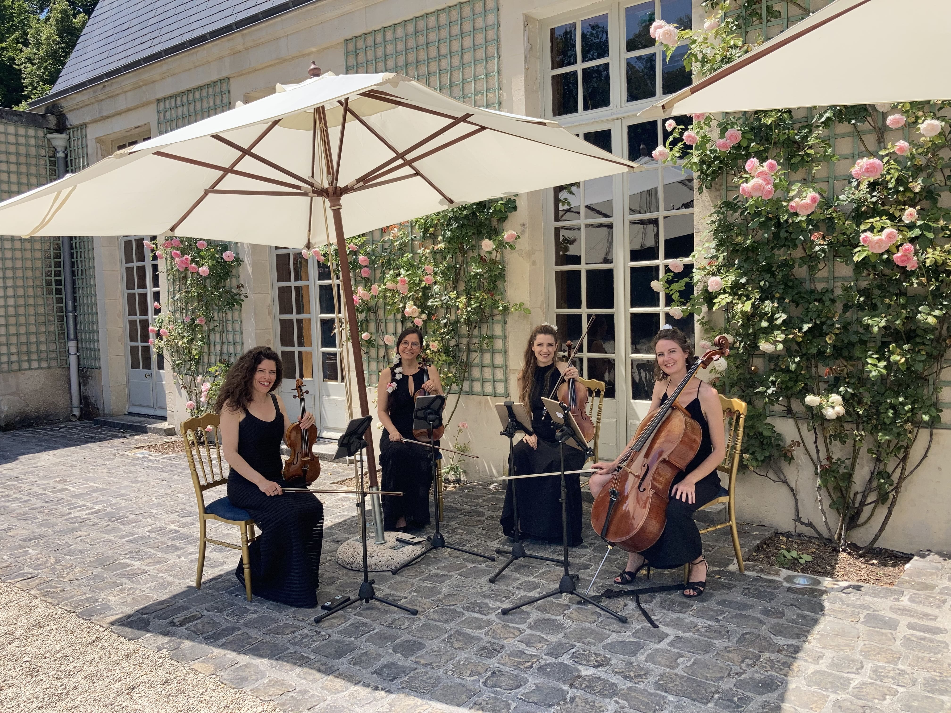 Four female musicians in black dresses seated outdoors under large white umbrellas, each holding a string instrument in front of a building with pink roses on trellises.