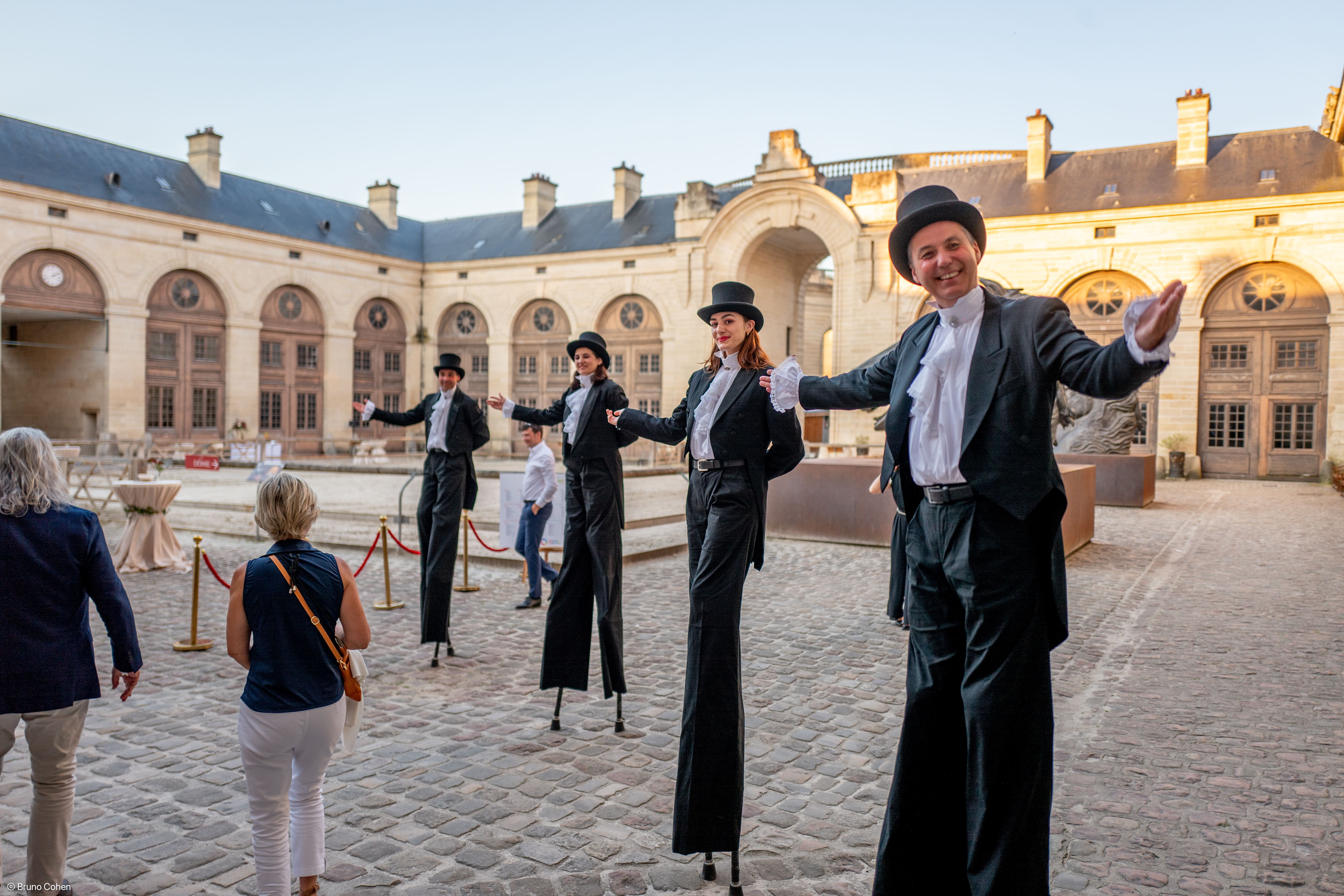 Four performers on stilts dressed in black suits, white shirts with ruffles, and top hats greeting visitors in a historic courtyard with stone cobblestones.