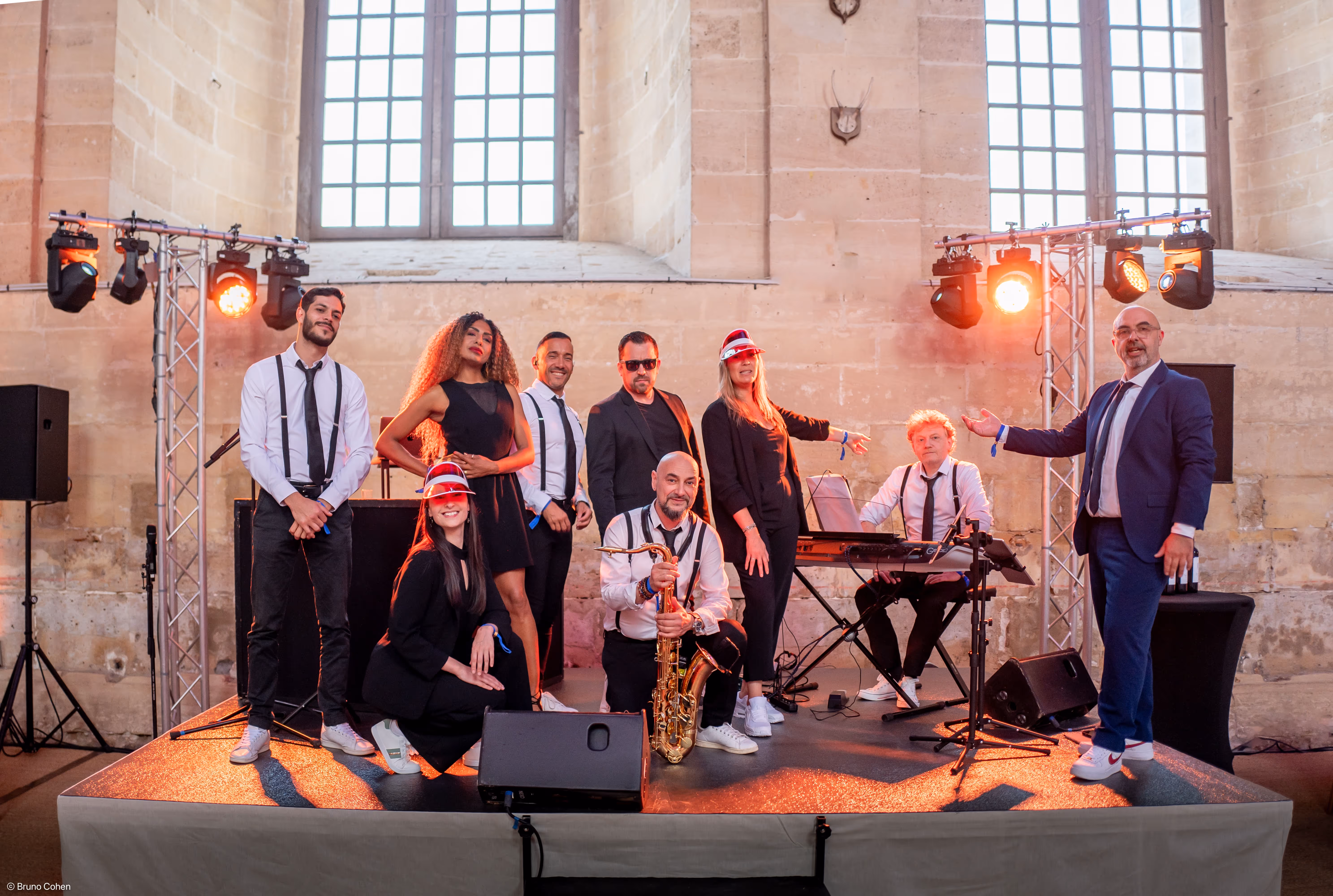 A band of eight musicians posing on a stage in front of large windows, with one holding a saxophone and another sitting at a keyboard, dressed in formal and semi-formal attire.