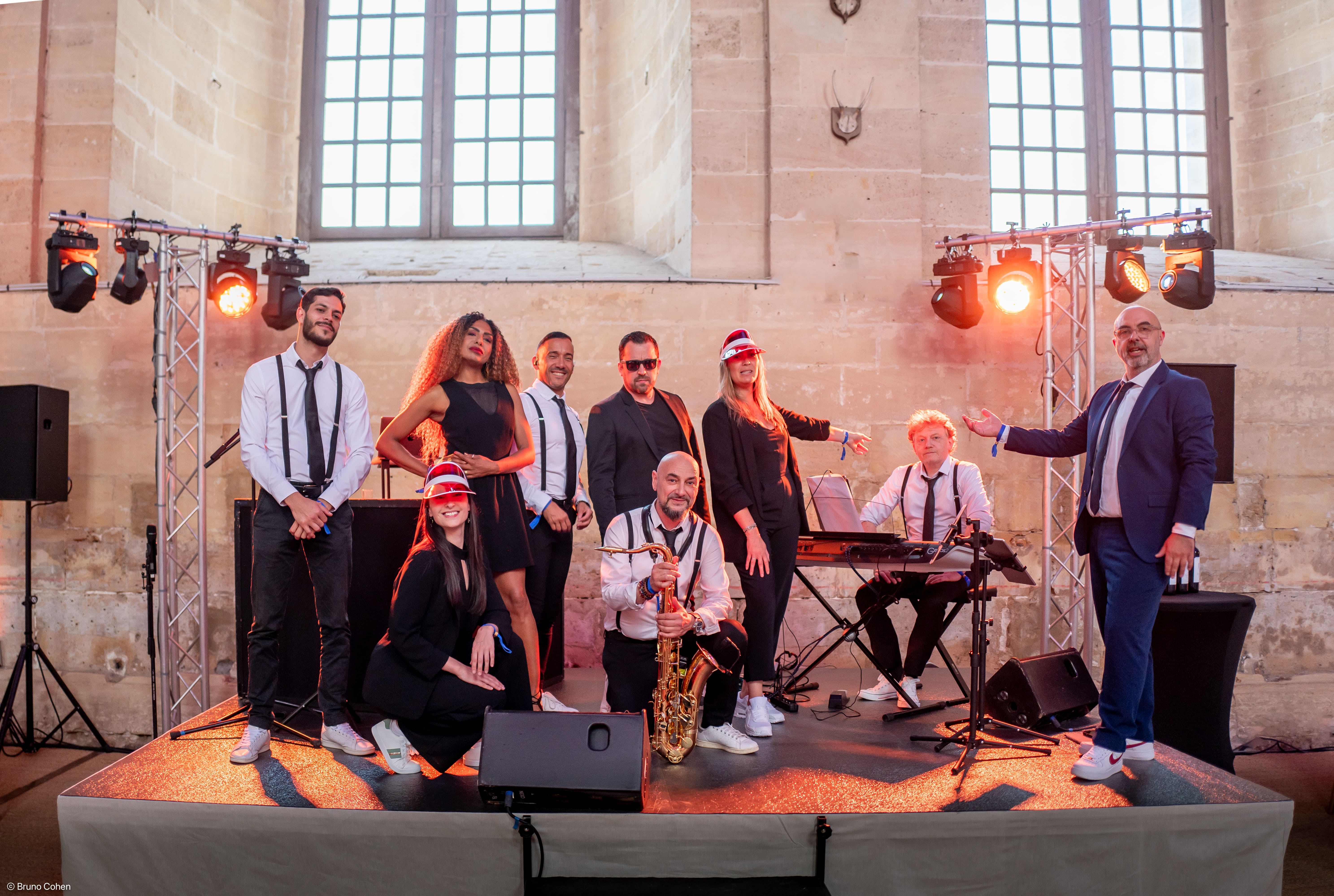 A band of eight musicians posing on a stage in front of large windows, with one holding a saxophone and another sitting at a keyboard, dressed in formal and semi-formal attire.