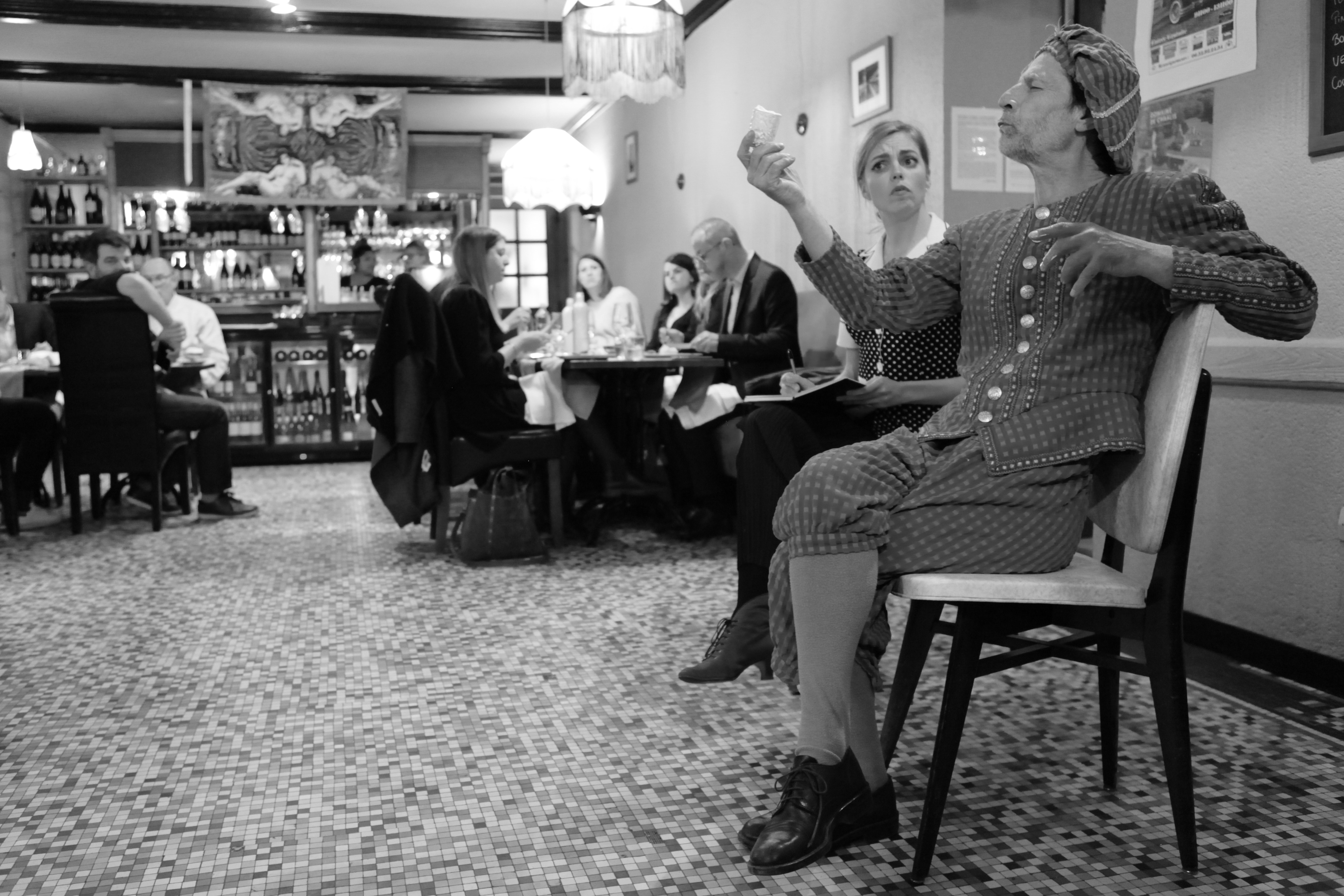 Man in historical costume sitting on a chair and gesturing with a small cup in a restaurant with seated diners in the background.