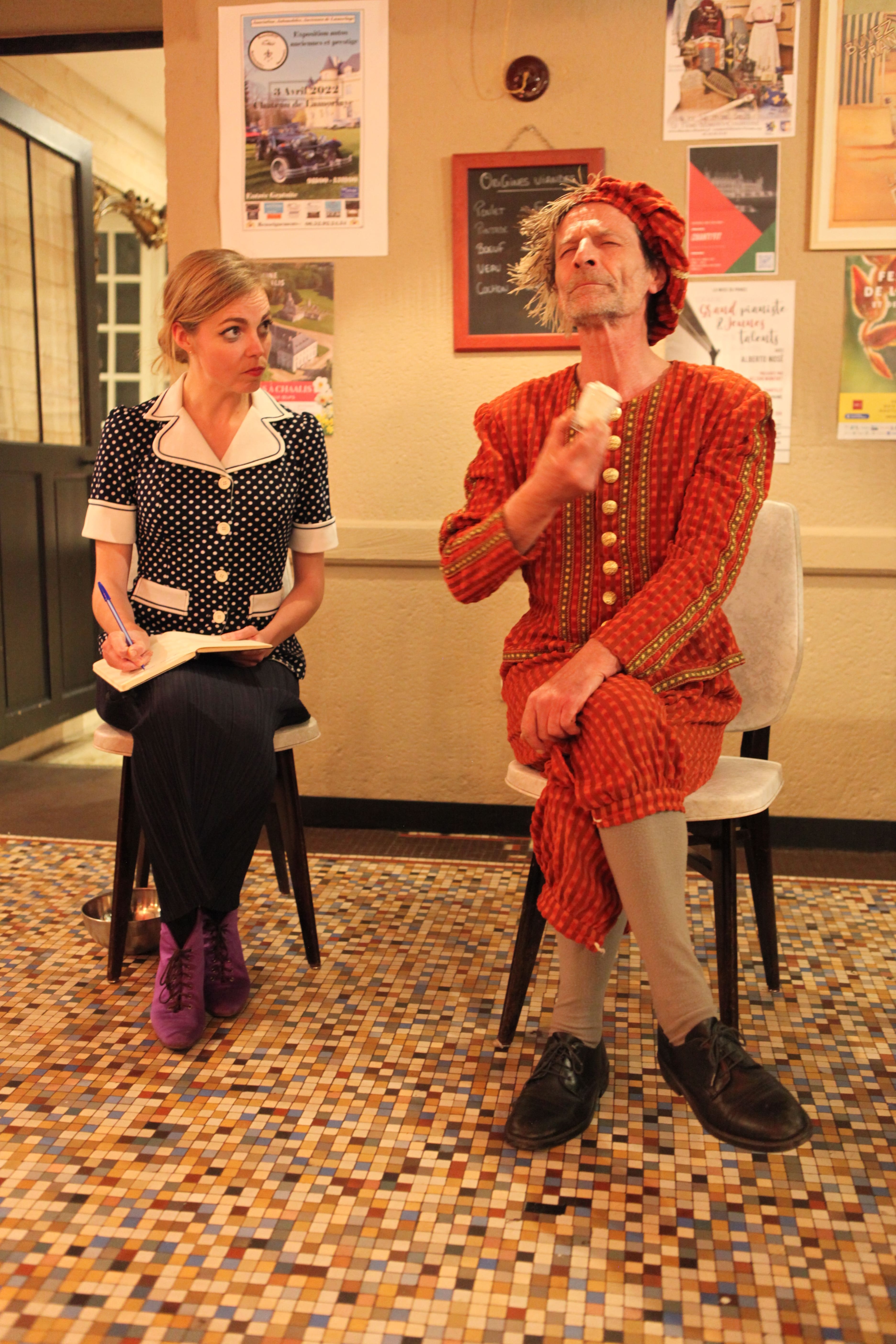 Man in red medieval costume sitting with legs crossed while a woman in a polka dot blouse writes in a notebook beside him.