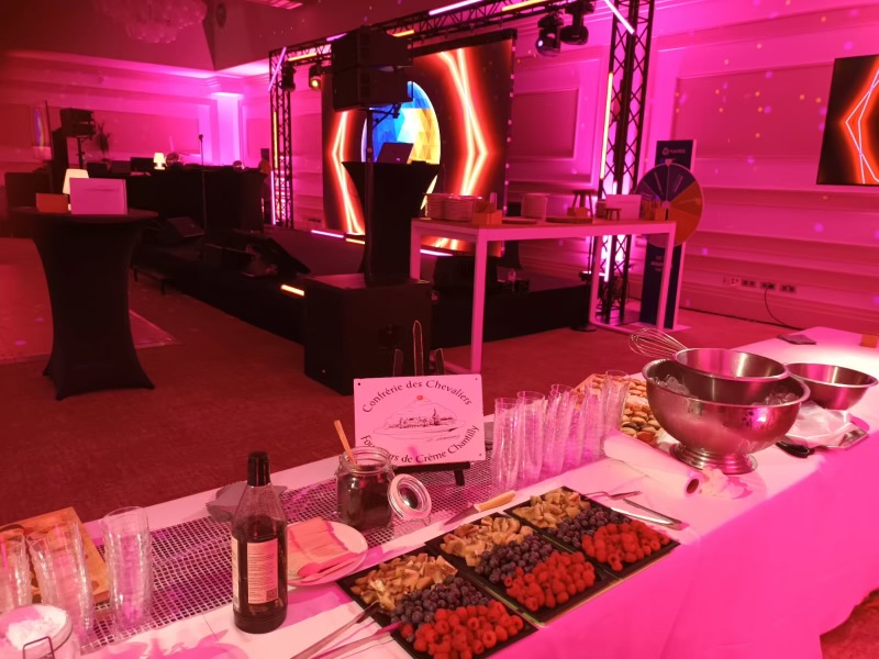 Event setup with a dessert table featuring bowls, trays of assorted berries, glasses, and a sign reading 'Confrérie des Chevaliers Foofeurs de Crème Chantilly' under pink lighting.