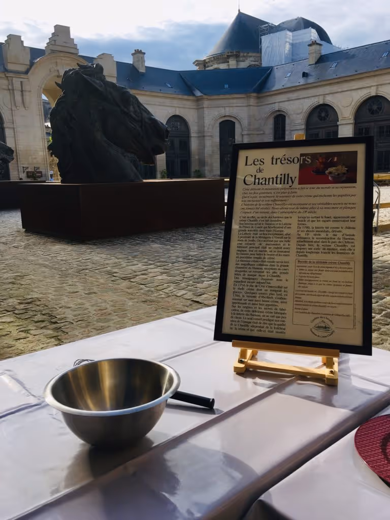 Outdoor courtyard with a large horse head sculpture, a table with a metal bowl and whisk, and an informational sign about Les trésors de Chantilly in French.