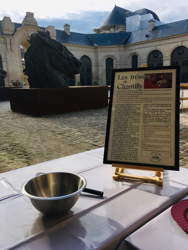 Outdoor courtyard with a large horse head sculpture, a table with a metal bowl and whisk, and an informational sign about Les trésors de Chantilly in French.