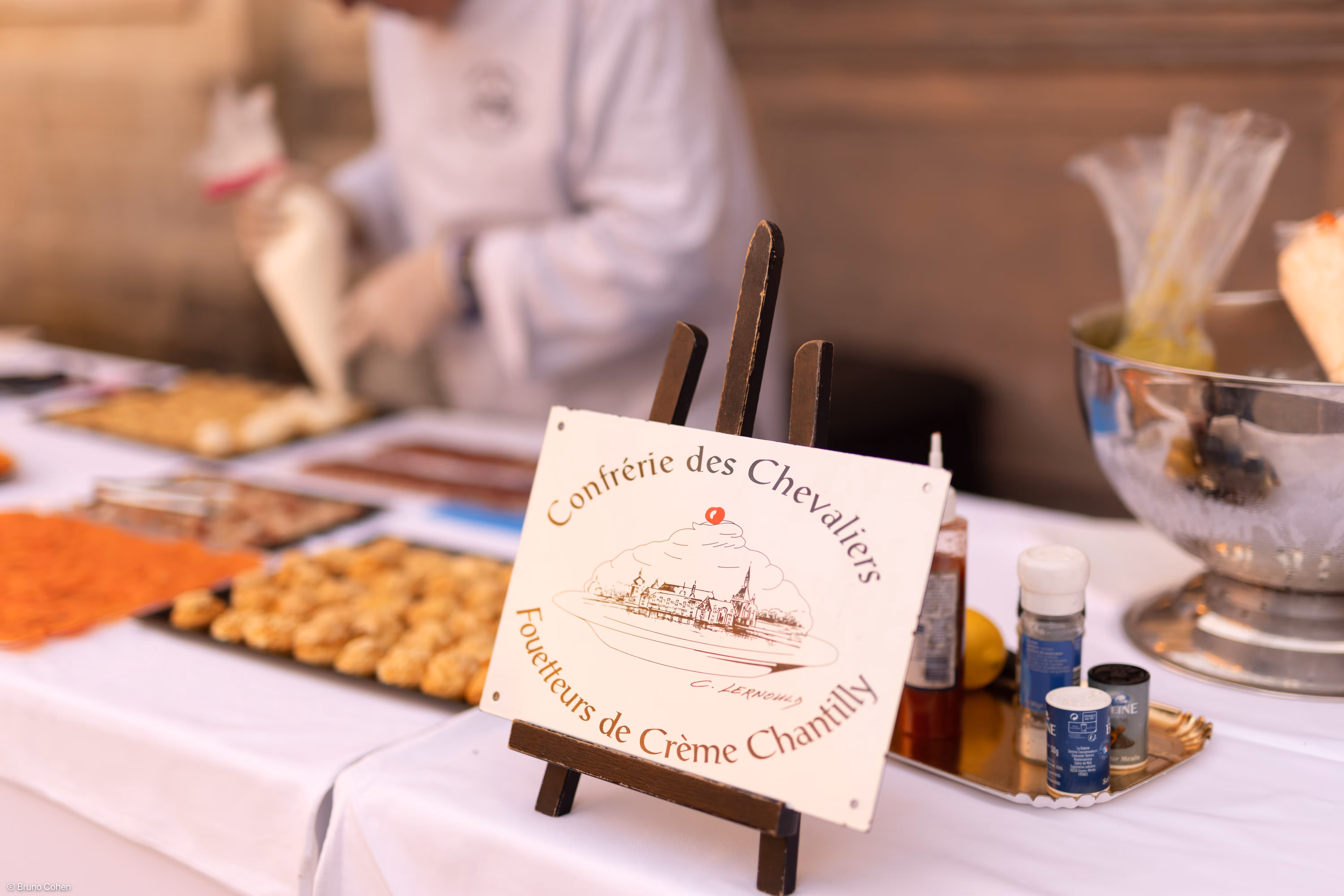 Sign on an easel reading 'Confrérie des Chevaliers Fouetteurs de Crème Chantilly' with a blurred background of a person piping cream and trays of pastries.