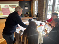 Four people around a round table tasting wine during a speed tasting event in a sunlit room with a projection screen showing a Speed Tasting Challenge description.
