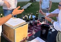 Group of men outdoors around a table with wine bottles, glasses, and documents, appearing to conduct a wine tasting near a historic building.