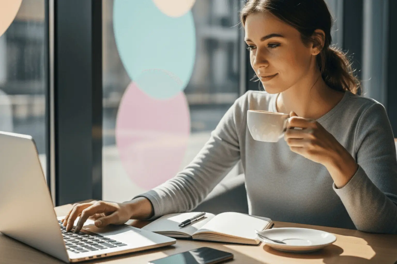 Junge Frau sitzt entspannt im Café mit Laptop und Smartphone, arbeitet an ihrer Online-Strategie bei natürlichem Tageslicht