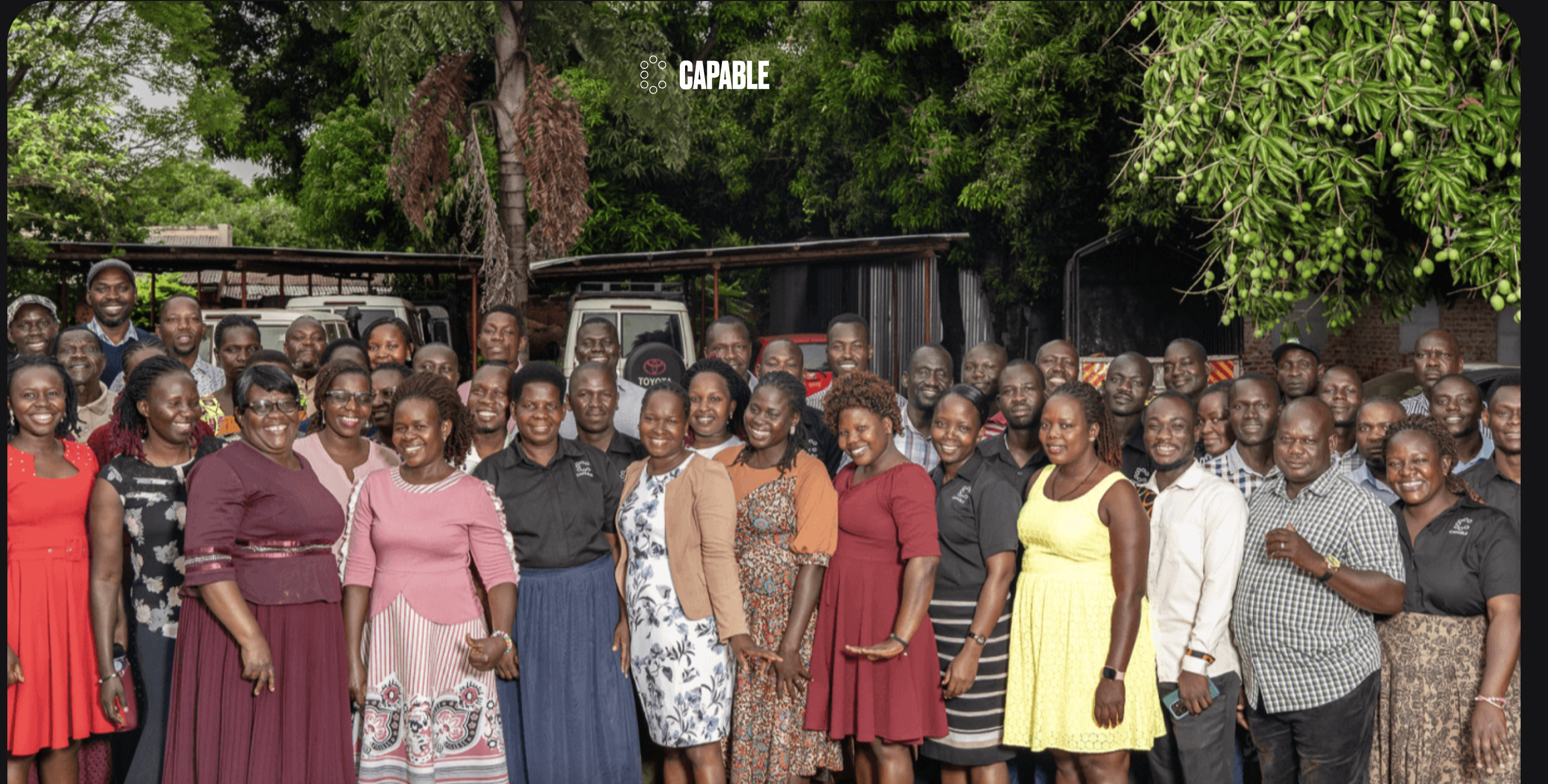 Large group photo of the diverse staff team for the CAPABLE organization, primarily featuring African men and women smiling outdoors in front of their vehicles and work structures.