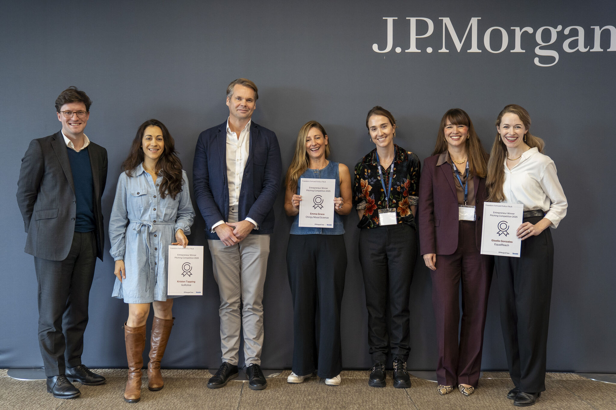 A group photo of seven individuals (four women and three men), standing in a line in front of a dark blue-gray wall with the J.P. Morgan logo. Four of the women are holding award certificates or papers.