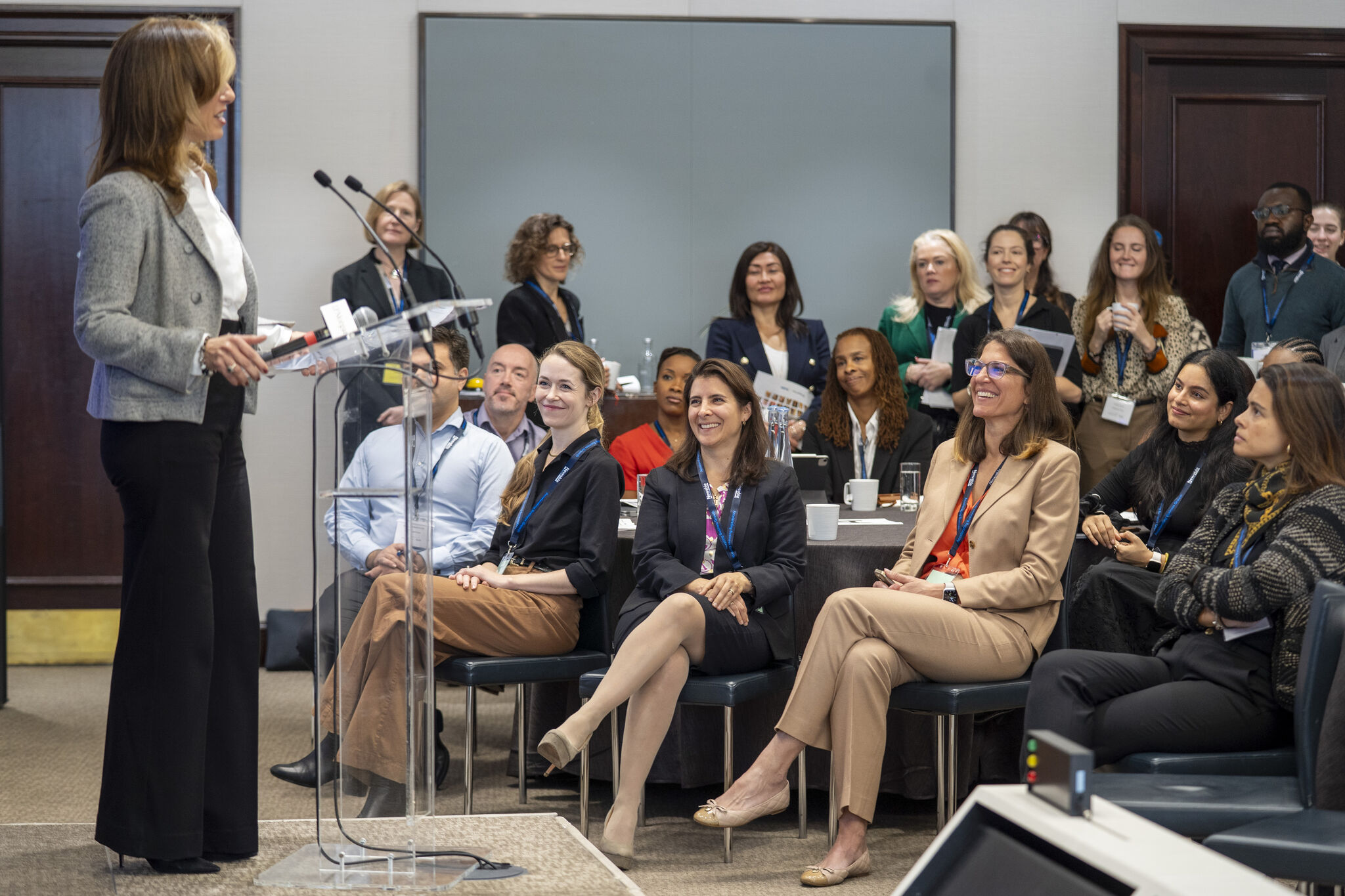 A female speaker in a gray jacket addresses an engaged audience from behind a clear podium during a presentation. The audience is seated, with several attendees smiling, listening, and looking at the speaker.