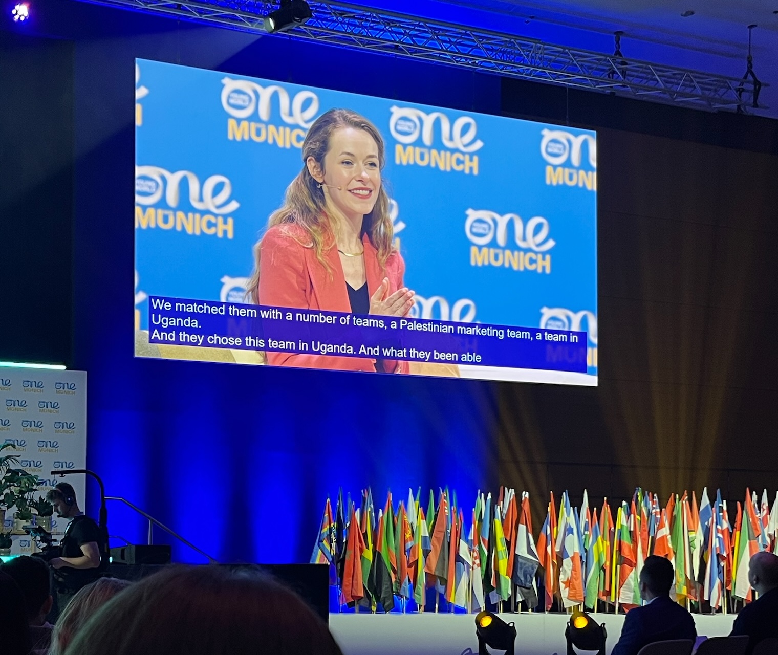 A speaker, Giselle, at a conference or One Young World summit in Munich. She is pictured on a large video screen wearing a coral blazer and speaking. The subtitle text visible is: "We matched them with a number of teams, a Palestinian marketing team, a team in Uganda. And they chose this team in Uganda. And what they been able..." The stage below the screen is decorated with a display of numerous national flags.