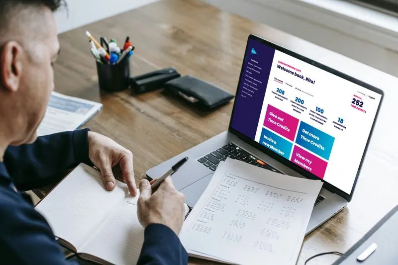 Man in a blue shirt is working at a wooden desk, writing in a notebook while looking at a laptop. The laptop displays a "Time Enterprise Cafe" dashboard with a welcome message for "Rita," showing Time Credit balances and action buttons like "Give out Time Credits" and "View my Members."