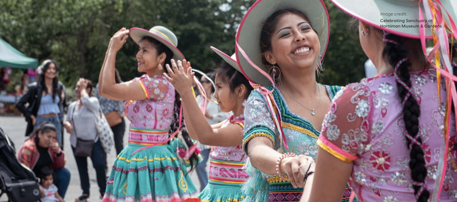 Several women and girls in colorful traditional dresses and wide-brimmed hats dance outdoors at a cultural celebration, smiling and holding hands.