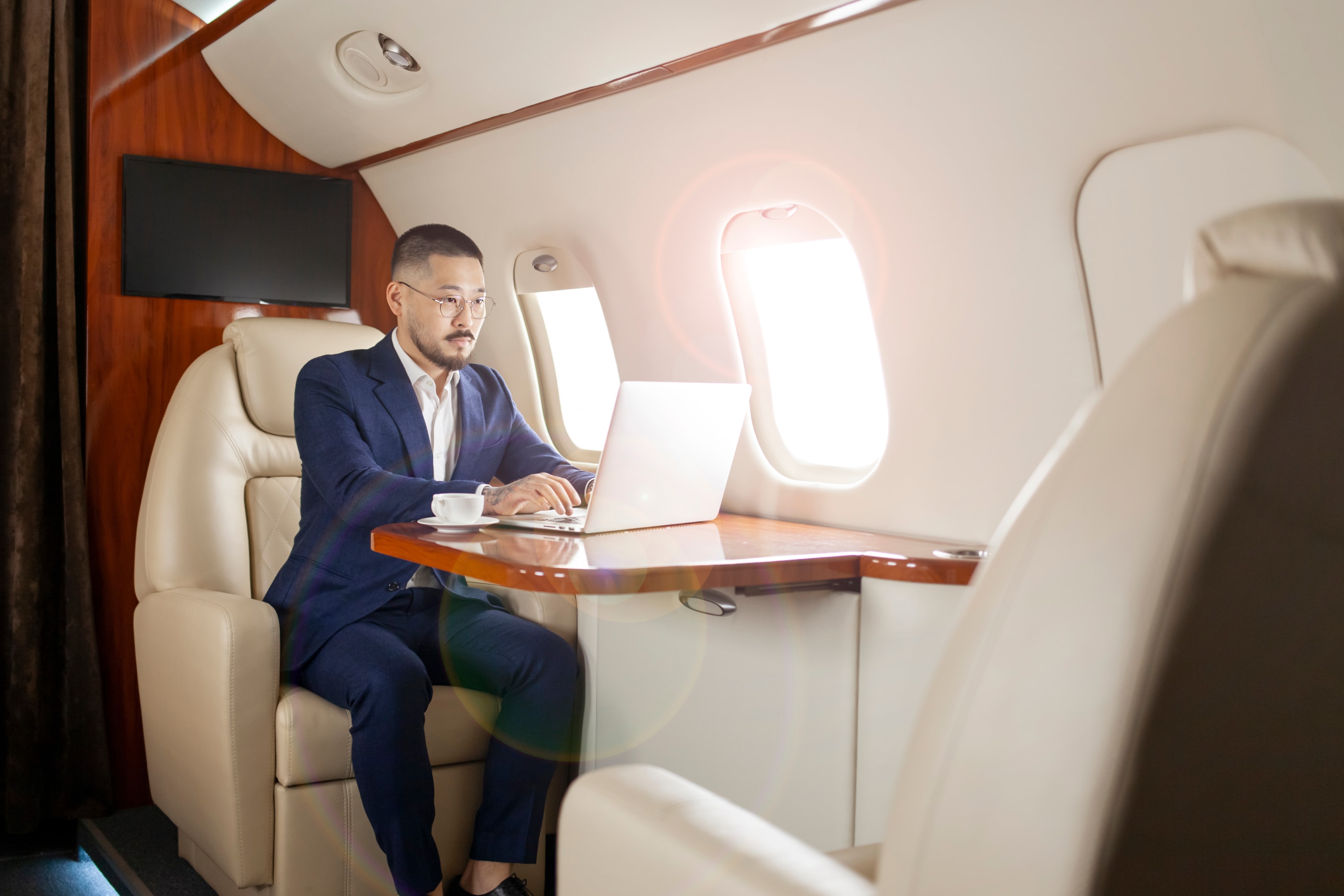 businessman in suit and glasses sits in private jet and types on laptop
