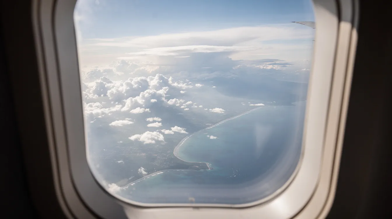 The image shows a view from an airplane window, featuring a blanket of fluffy clouds below and a distant coastline visible on the horizon. This scenic aerial perspective captures the essence of air travel, perfect for those planning their next trip or looking for the best time to purchase plane tickets.